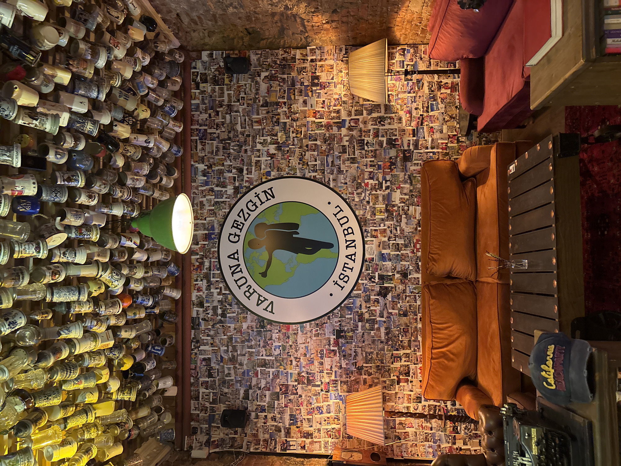 The image depicts the interior of a dimly lit, cozy establishment, likely a bar or restaurant, with a distinctly eclectic and personal decor. The walls are densely covered with a collage of photographs, creating a textured and vibrant backdrop. Prominently displayed in the center of the image is a circular sign with an image of the Earth and a silhouette of a person, surrounded by text that appears to be in Turkish, possibly reading "GEZGİN BUL. İSTANBUL," which translates to "Traveler Find. Istanbul." This suggests a travel-themed venue or a place frequented by travelers.
In the foreground, a collection of ceramic mugs, possibly beer steins, are mounted on the wall, showcasing various designs and patterns. To the right, a plush orange sofa and a darker, tufted leather chair create a comfortable seating area. A wooden table with a dark, slatted surface is positioned in front of the sofa. On the table, a small vase with sprigs of dried flowers is visible. Nearby, a vintage typewriter sits on a shelf. A blue baseball cap with the embroidered text "California Beach Party" rests on the shelf next to the typewriter, adding a touch of casual Americana.
A green industrial-style lamp with a bright light fixture is suspended from the ceiling, illuminating a portion of the wall adorned with photos. Another lampshade, with a ribbed texture and an orange glow, is situated on a table behind the sofa, casting a warm light. The ceiling appears to be constructed from exposed brick, contributing to the rustic ambiance. The overall atmosphere is one of relaxed camaraderie and a celebration of travel and memories, as evidenced by the abundant photographs on the walls. The time of day is indeterminate, but the lighting suggests an evening or indoor setting.