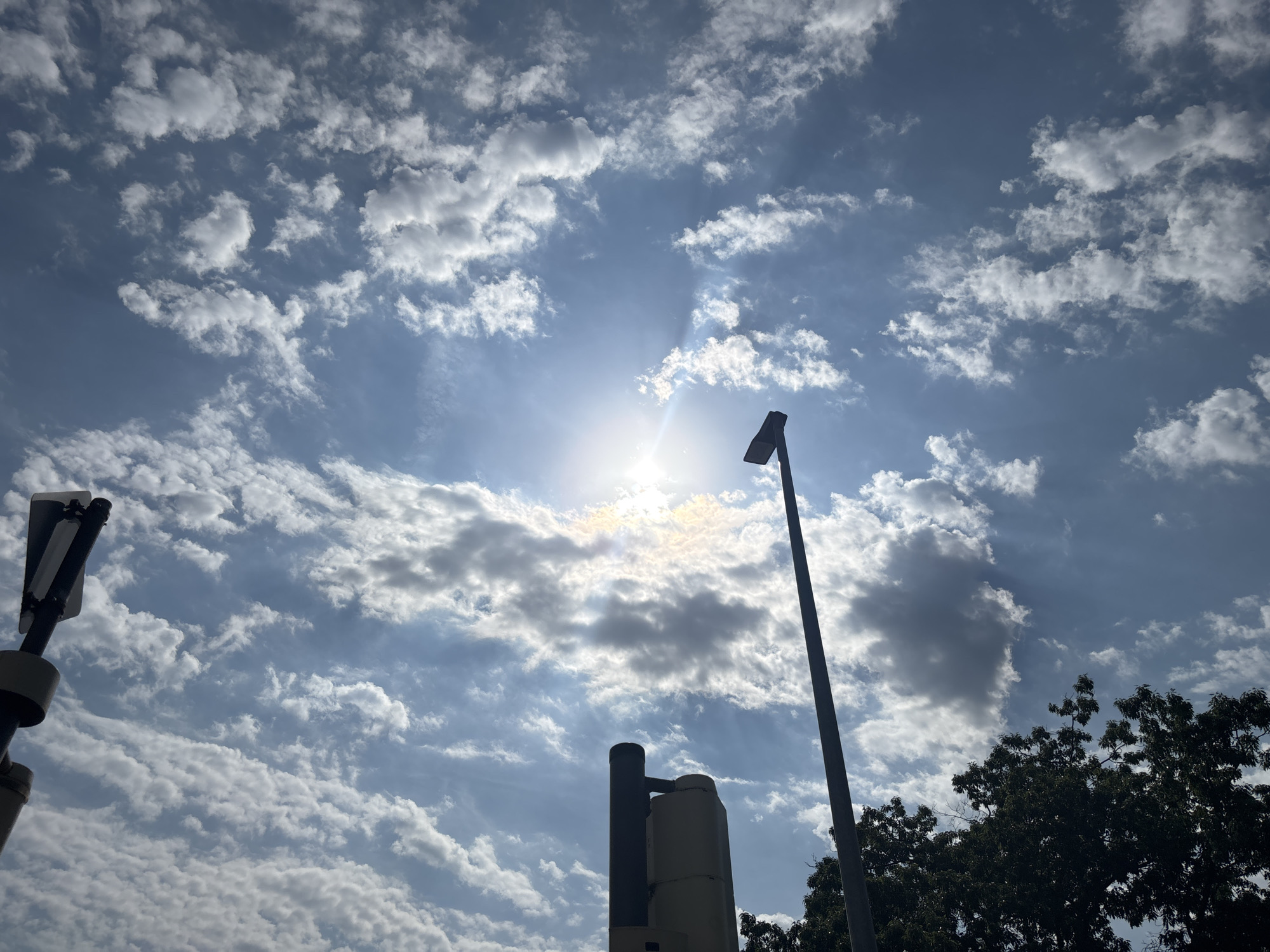 The image shows a bright, sunny day with a blue sky filled with scattered white and gray clouds. The sun is visible, shining brightly through some of the clouds, creating visible light rays. In the foreground, silhouetted against the sky, are various man-made structures and natural elements. On the left side, a traffic signal or a street signpost is partially visible. To the right of that, a tall, dark pole extends upwards, topped with a streetlamp. In the center of the image, a cylindrical structure, possibly part of a utility pole or a smaller, modern sculpture, is visible. Further to the right, the dark green foliage of a large tree is seen against the sky. The overall impression is of an urban or suburban outdoor setting, likely a street or park, on a partly cloudy, sunny day. There are no people visible in the image. The scene does not provide any specific visual cues that would allow for definitive identification of its location within the city of Celle, Germany.