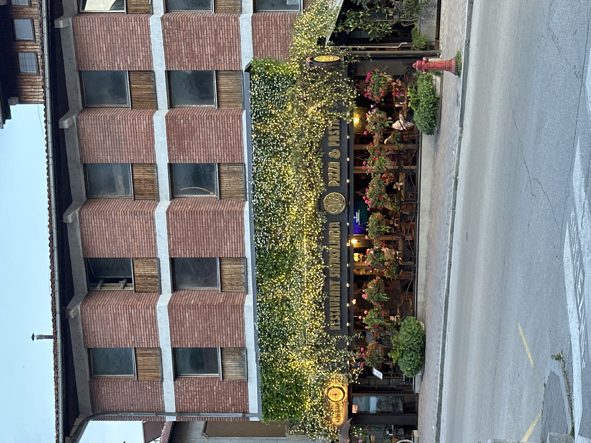 The image captures the exterior of "Restaurant Shisharkata Pizza & Pasta" in Veliko Tarnovo, Bulgaria. The restaurant is situated on the ground floor of a multi-story brick building. The facade is decorated with a lush green vine and numerous small, warm-toned lights, creating an inviting ambiance. Hanging flower baskets filled with pink and red blooms are displayed prominently above the entrance. The restaurant's name is visible on a dark sign above the windows, along with a clock and a circular emblem featuring what appears to be a laurel wreath. Through the large windows, patrons can be seen seated at tables. A red fire hydrant stands on the sidewalk in front of the establishment. The scene is set on a street, with a paved road and sidewalk visible in the foreground. The sky is overcast, suggesting it is either early morning or late afternoon.