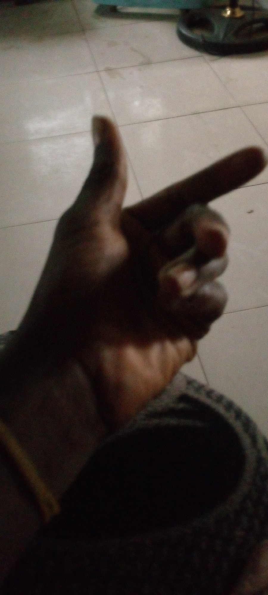 A partially visible human hand is shown against a light tiled floor. The hand is dark-skinned and positioned with the thumb up and index finger extended, resembling a gun. The hand extends from a dark fabric covering part of the lower portion of the image, likely clothing. A partial view of an unidentified dark-colored object sits on the floor in the background. Location is undetermined.