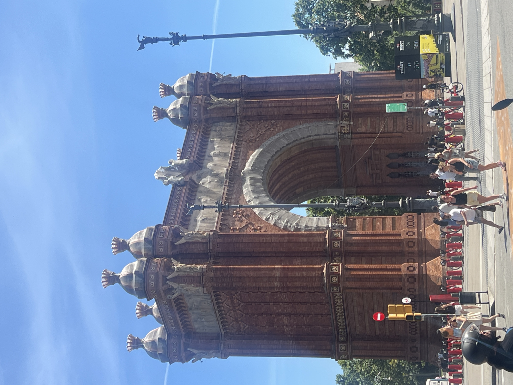 The Arc de Triomf in Barcelona, Spain, is captured in this frontal, eye-level shot on a clear, sunny day. The monument, constructed from red brick, dominates the frame with its arched opening and ornate detailing. Above the arch, several decorative cupolas topped with crowns are visible against a bright blue sky with faint contrails. Sculptural figures of winged beings adorn the sides of the archway. In the foreground, a busy street scene unfolds. Pedestrians walk across a crosswalk, some in casual summer attire. Traffic signals and a "no entry" sign are present on the left. A dark-colored motorcycle helmet is positioned at the bottom left of the frame, suggesting the perspective of a rider. In the middle ground, a line of red and silver bollards separates pedestrian areas from the street. Behind the bollards, several bicycles are parked. A Tissot advertising board is visible on the right, displaying timers. Lush green trees frame the monument on the right side.