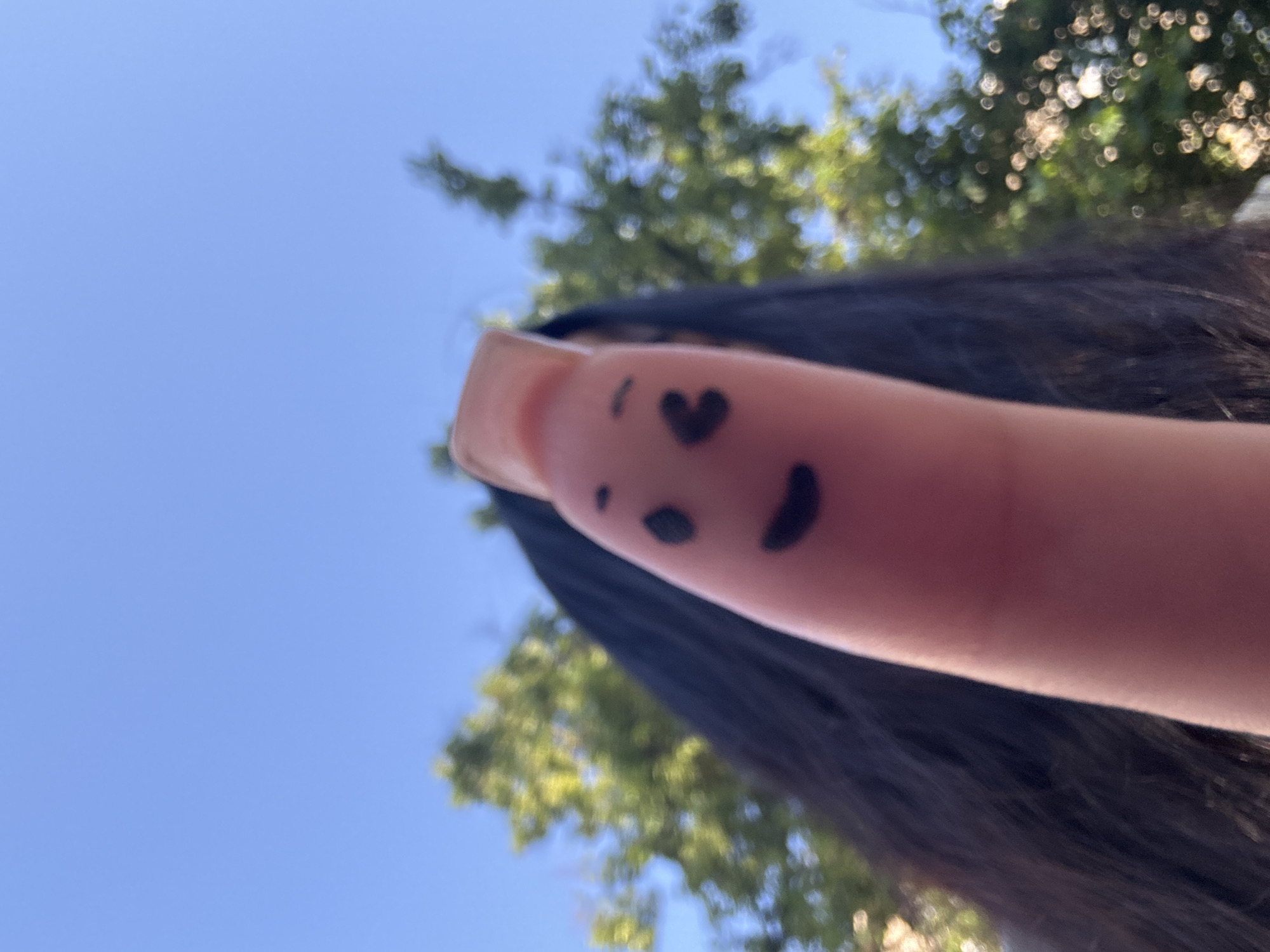 A close-up, low-angle shot captures a thumb with a smiley face drawn on it. The thumb is positioned in the foreground, with the face created using black marker. The face has two dots for eyes and a curved line for a mouth. Behind the thumb, a person's dark hair is visible, and beyond that, blurred green trees are seen against a clear blue sky. The image appears to be taken outdoors. The location of Novosibirsk, Russia, is not indicated by any visual elements in the image.
