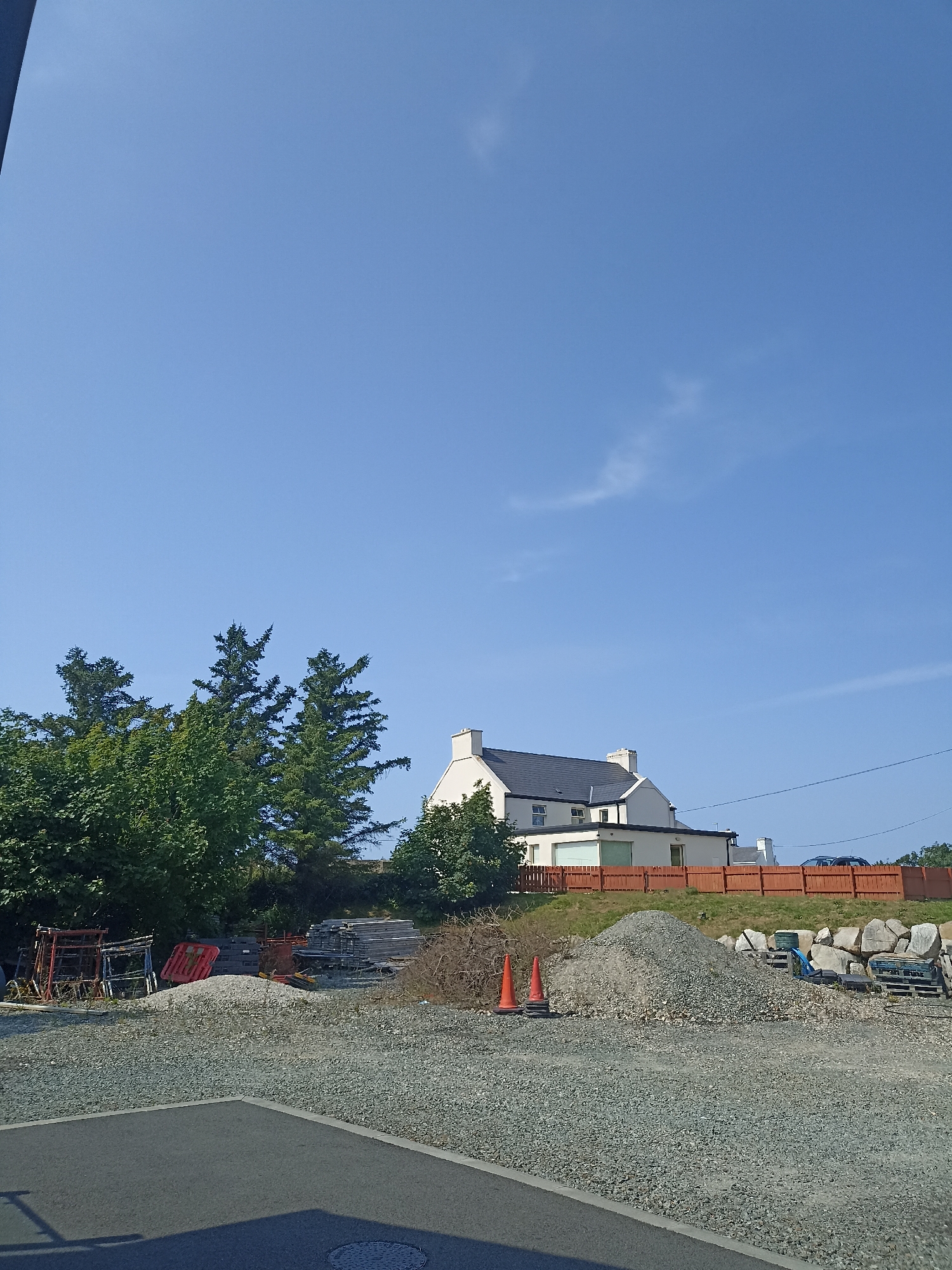 The image depicts a construction or storage yard in Doirí Beaga, Ireland, on a clear, sunny day. A residential building with white walls and a dark roof is visible in the background, situated behind a wooden fence. The foreground is dominated by a gravel surface, with piles of stones and construction materials scattered throughout. Several orange traffic cones and stacks of wooden pallets are present. Lush green trees and foliage line the left side of the image, contrasting with the man-made structures. The sky is a vibrant blue with a few wispy clouds. No people are visible in the scene.