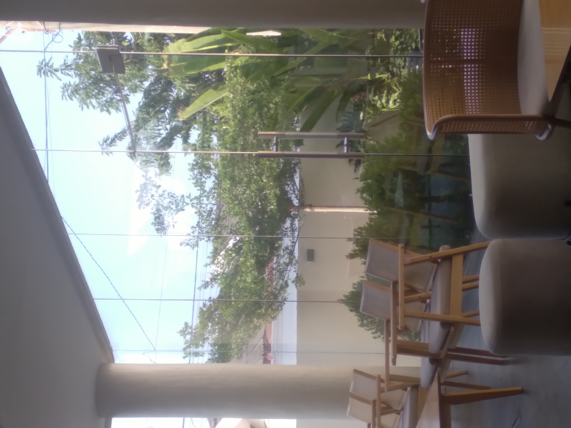 The image depicts a view from inside a building, likely a café or restaurant, in Lhokseumawe, Indonesia. The foreground features furniture including woven chairs and upholstered seating. A large window provides a view of lush green foliage, trees, and a building exterior. The reflections on the window suggest a bright, possibly sunny, day. The scene appears calm and relaxed, with no visible people or active interactions.