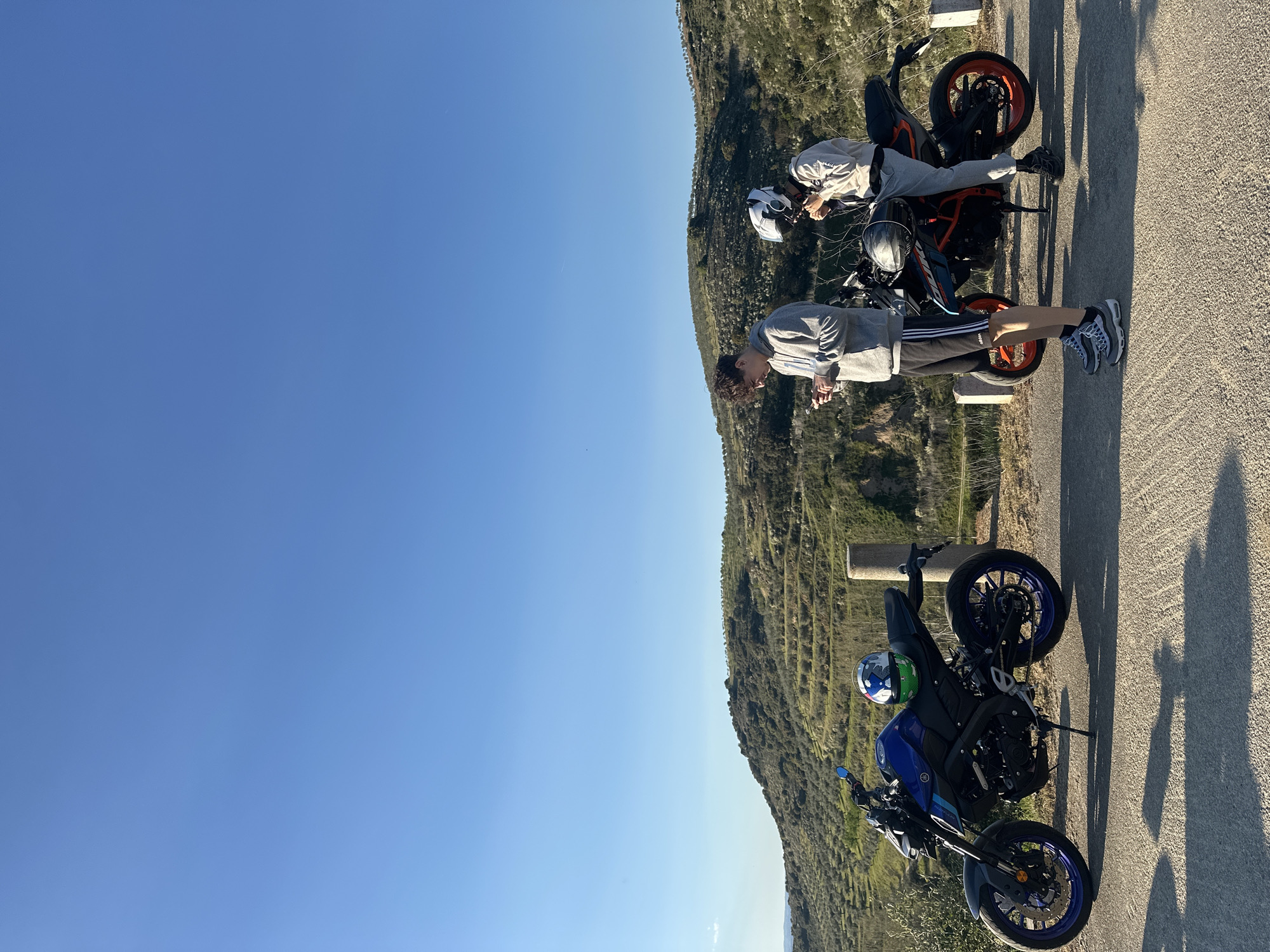 Two young men stand on a roadside overlooking a scenic landscape near Mirandela, Portugal. One is near a blue Yamaha MT-03 motorcycle, wearing a grey hoodie and black shorts with white stripes, holding something in his hands. The other is near an orange and black KTM motorcycle, wearing a light grey outfit. Both motorcycles have helmets placed on them. The background shows a hillside and a clear blue sky.
