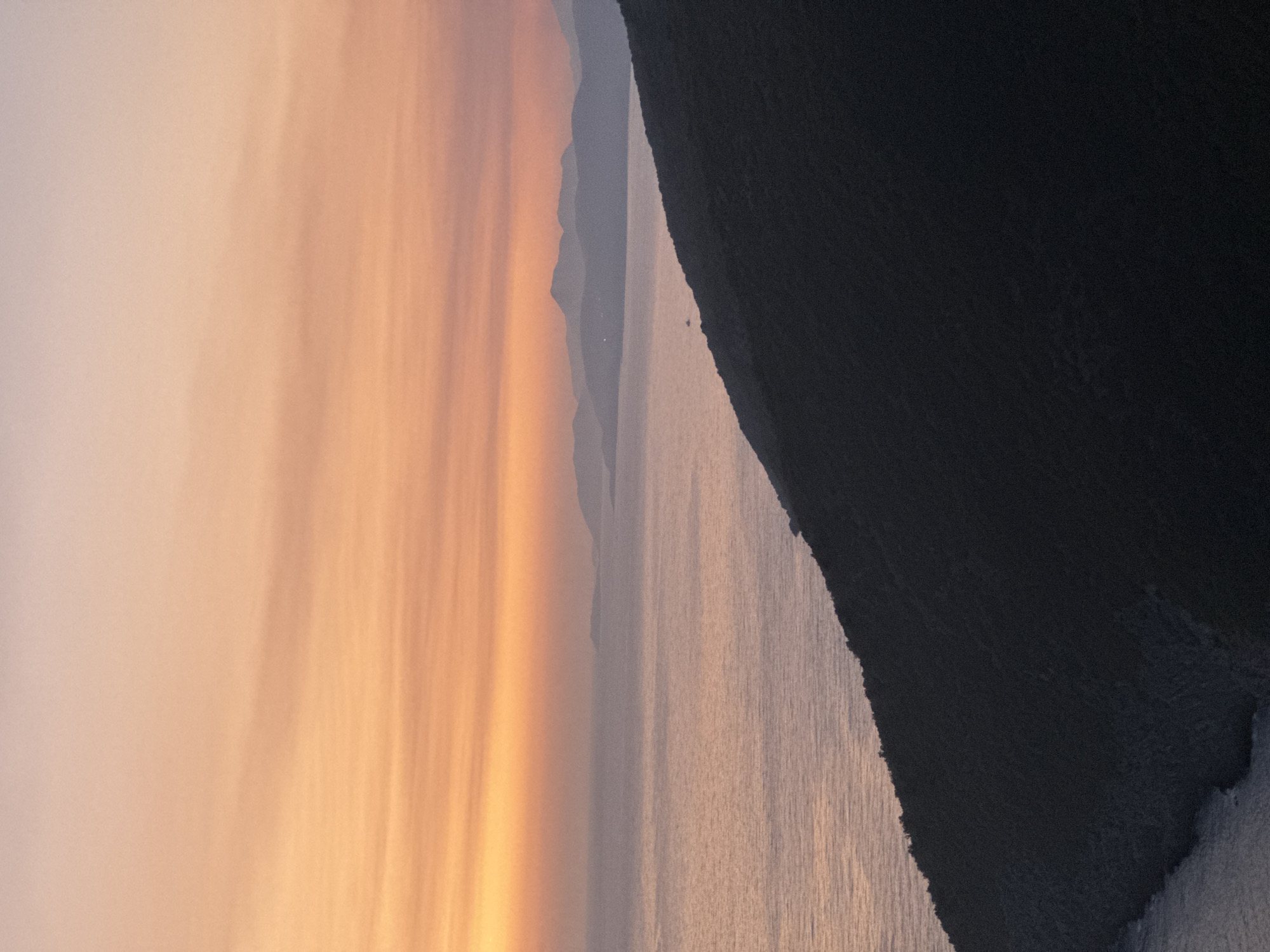 The image depicts a coastal landscape at sunset, likely near Zagora, Montenegro. A dark, densely vegetated hillside slopes diagonally from the foreground into the middle ground, meeting the calm, subtly rippling water of the Adriatic Sea. The sea extends to the horizon where a series of silhouetted mountains, their forms softened by distance and atmospheric haze, rise against the sky. The sky itself is a gradient of warm colors, transitioning from a soft peach and orange near the horizon to a pale pink and lavender at the top of the frame. Thin, wispy clouds are visible, adding texture to the sunset. A small, dark boat is discernible on the water in the mid-distance. The overall impression is one of serene natural beauty and tranquility.
