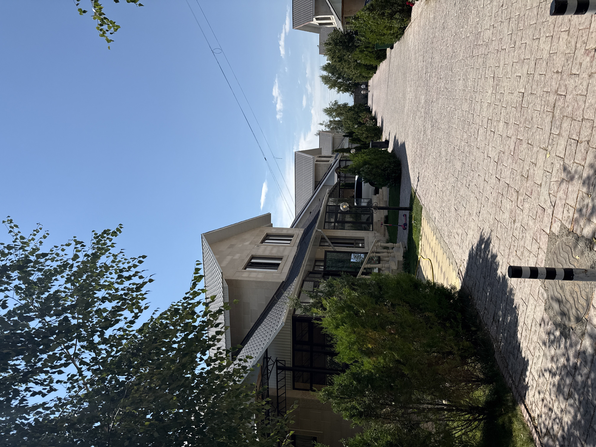 A street lined with modern, two-story houses and green trees under a clear blue sky. The houses are made of light-colored stone and have dark tiled roofs. A cobblestone path runs down the center of the street, with green lawns and manicured bushes on either side. Two people are walking away from the camera in the distance. The scene appears to be in a well-maintained residential area.