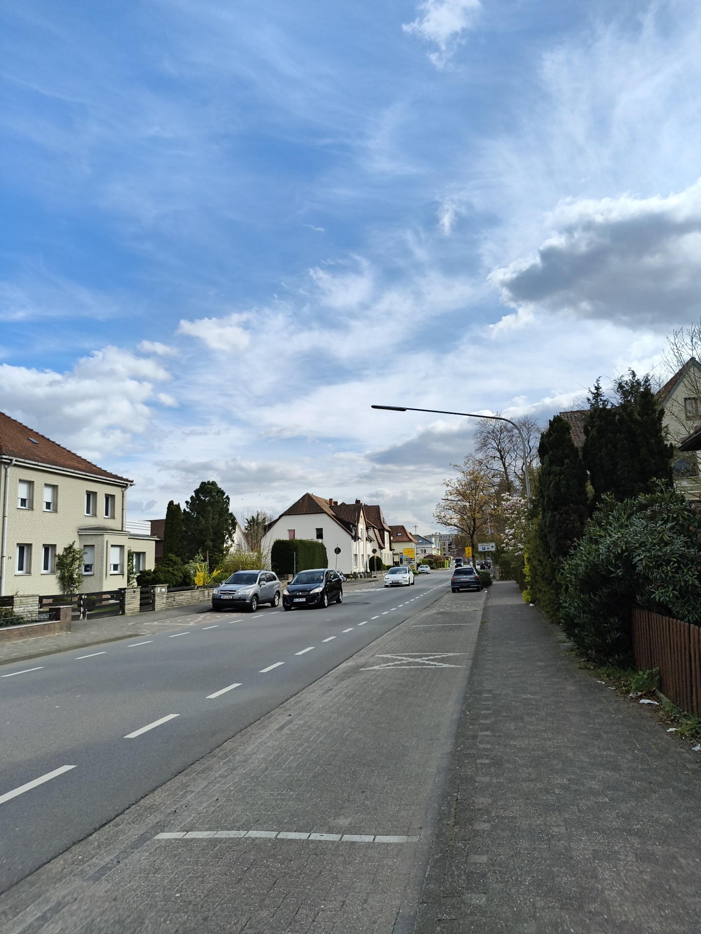 The image depicts a street scene in Diepholz, Germany. The view is along a residential street with houses lining both sides. A two-lane road runs through the middle of the frame, with white lane markings. Several cars are visible on the road, including a silver SUV and a dark-colored sedan parked on the left side, facing the camera. Further down the street, additional cars are driving in both directions. A sidewalk borders the road on the right, paved with grey bricks. A wooden fence and various bushes and trees separate the sidewalk from the properties on that side. A street light extends over the road from the right side. The sky is partly cloudy, with a mix of blue sky and white clouds. The buildings are typical for a European residential area.