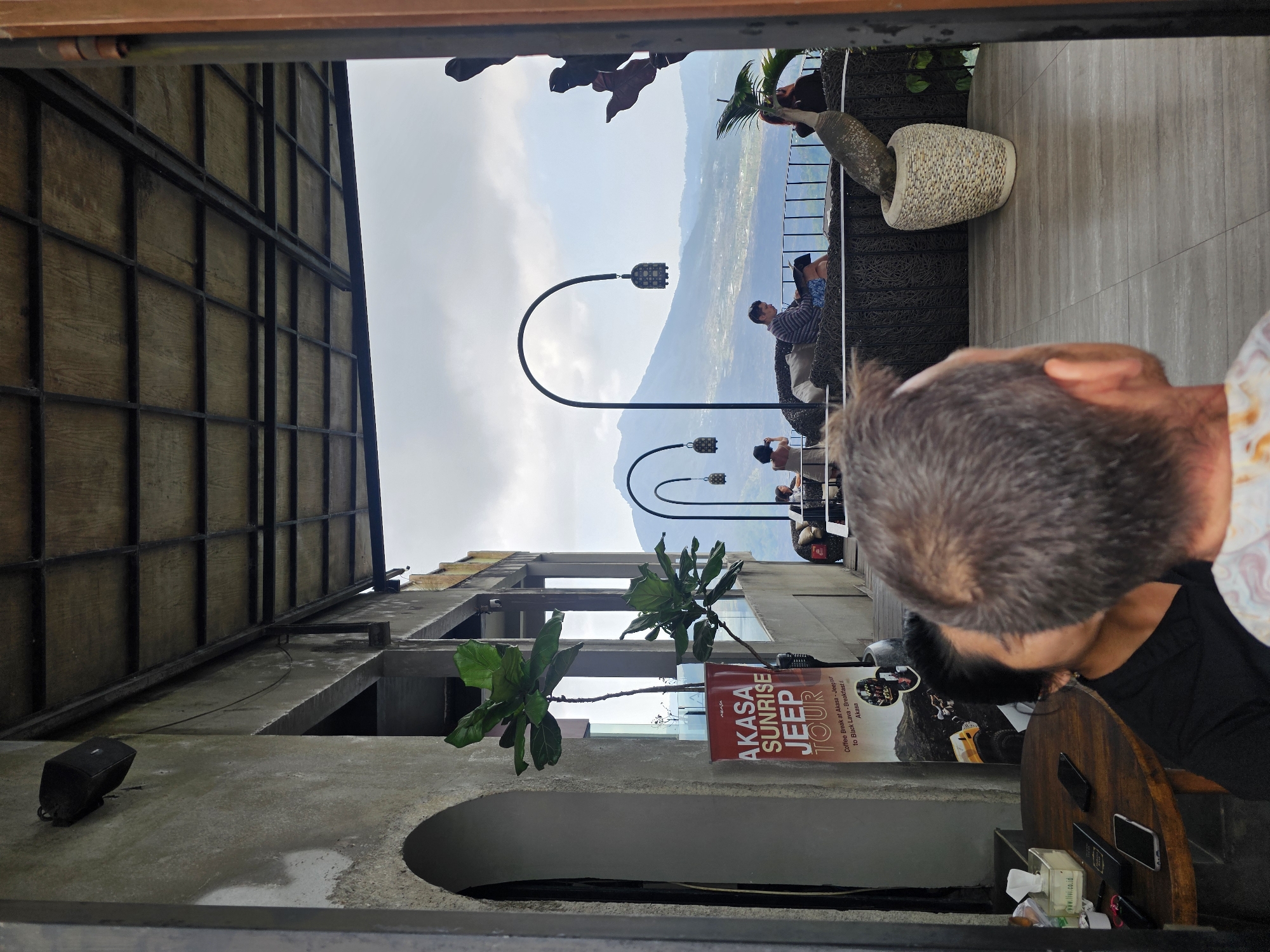 This is a medium shot of an outdoor seating area at a cafe in Kintamani, Indonesia, with a panoramic view of a mountainous landscape. In the foreground, a man with graying hair is looking away from the camera, his head and shoulders visible. To his left and slightly in front of him, a woman with dark hair, wearing a black shirt, is partially visible. A wooden table is to the left of the couple, with several mobile phones and a tissue box on it.
Behind the foreground figures, the cafe features a covered seating area with wicker furniture, where several patrons are seated and conversing. One man is reading a book while sitting on a wicker sofa. Beyond the seating area, a railing separates the cafe from a steep drop, offering a stunning view of a valley with scattered buildings and lush vegetation, leading to a mountain range under a cloudy sky. Decorative lamps are suspended from curved poles along the railing.
To the left of the seating area, there's a concrete wall with an arched opening and a large potted plant with broad leaves. A red banner advertising "AKASA SUNRISE JEEP TOUR" is prominently displayed near the plant. The architecture of the cafe is characterized by exposed concrete and a metal-framed roof with wooden panels. The overall atmosphere suggests a relaxed setting for enjoying the scenic beauty of Kintamani.