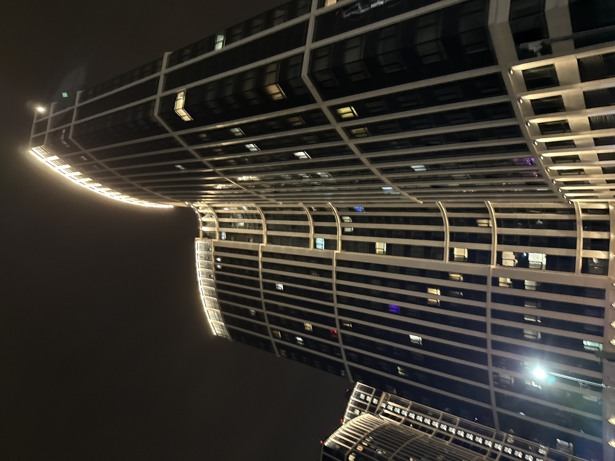 A low-angle, nighttime shot captures two illuminated high-rise buildings in Khimki, Russia. The structures are characterized by a grid-like facade of windows and horizontal beams. The upper building exhibits a curved edge illuminated with a strip of light. Various window lights are visible. The sky is dark, adding to the scene's contrast. A lower building with a curved roof and grid-like windows is visible at the bottom of the image. A bright light source is visible near the lower building, casting a glare in the image.