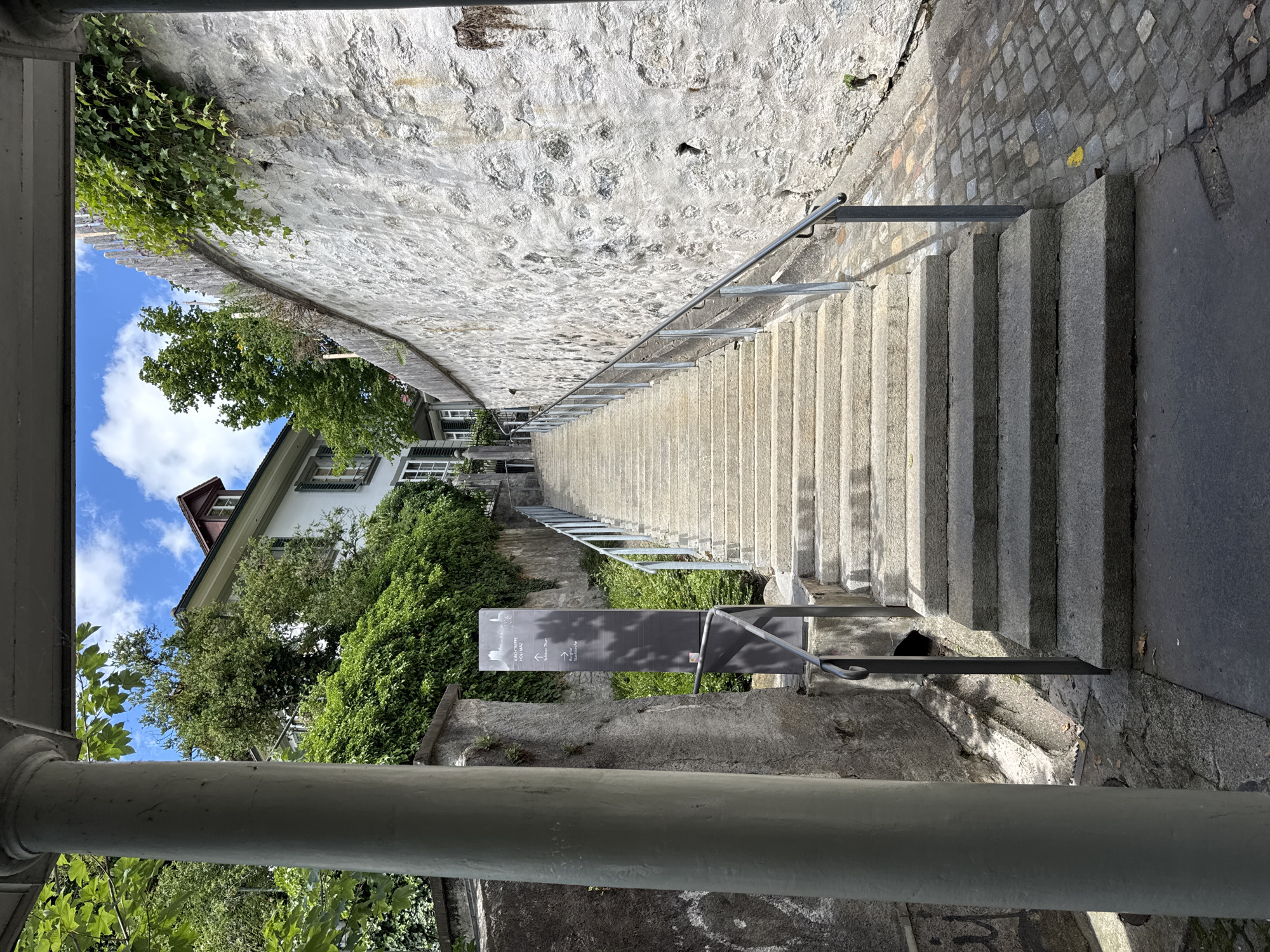 This is a vertical, eye-level, full-shot photograph taken from a sheltered pathway, looking up a wide, stone staircase leading towards a building in Thun, Switzerland. The staircase has metal railings on both sides and is bordered by a rough, textured stone wall on the right, which is partially covered by ivy. On the left, a large, fluted, grey column is visible in the foreground, partially obscuring the view. Adjacent to the staircase on the left is a grey concrete wall with some graffiti. A black directional sign with white text and graphics is positioned near the bottom of the staircase on the left. The building at the top of the stairs is white with a reddish-brown roof and green shutters. Lush green trees and foliage surround the building and climb the walls. The sky is blue with scattered white clouds. The pathway next to the stairs is paved with cobblestones.