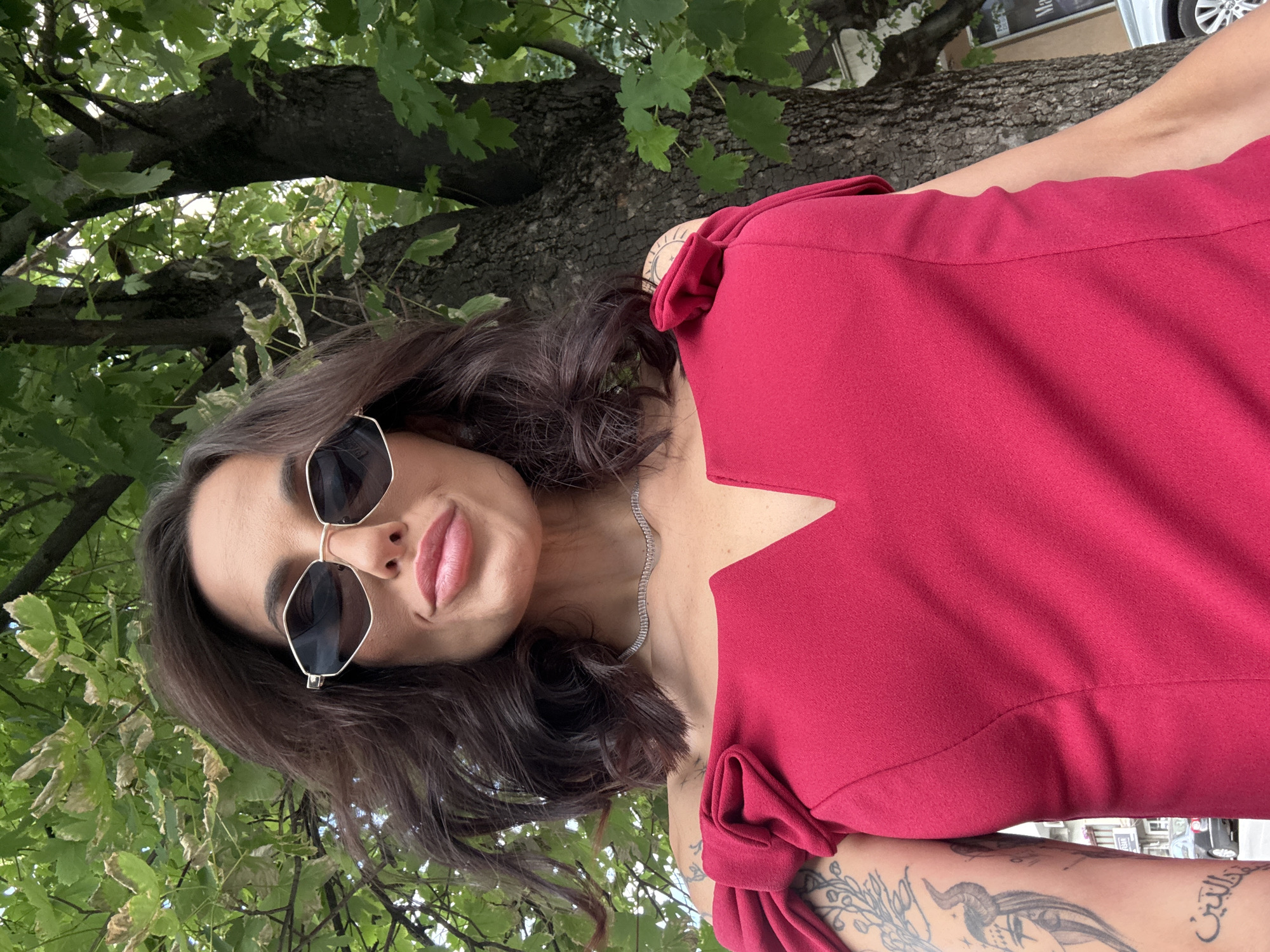 A woman is pictured from a low angle against a backdrop of foliage. The woman is wearing sunglasses and a red dress with a geometric neckline. She has dark hair, a necklace, and tattoos on her arms. The foliage appears to be from a tree. The location is Pleven, Bulgaria.