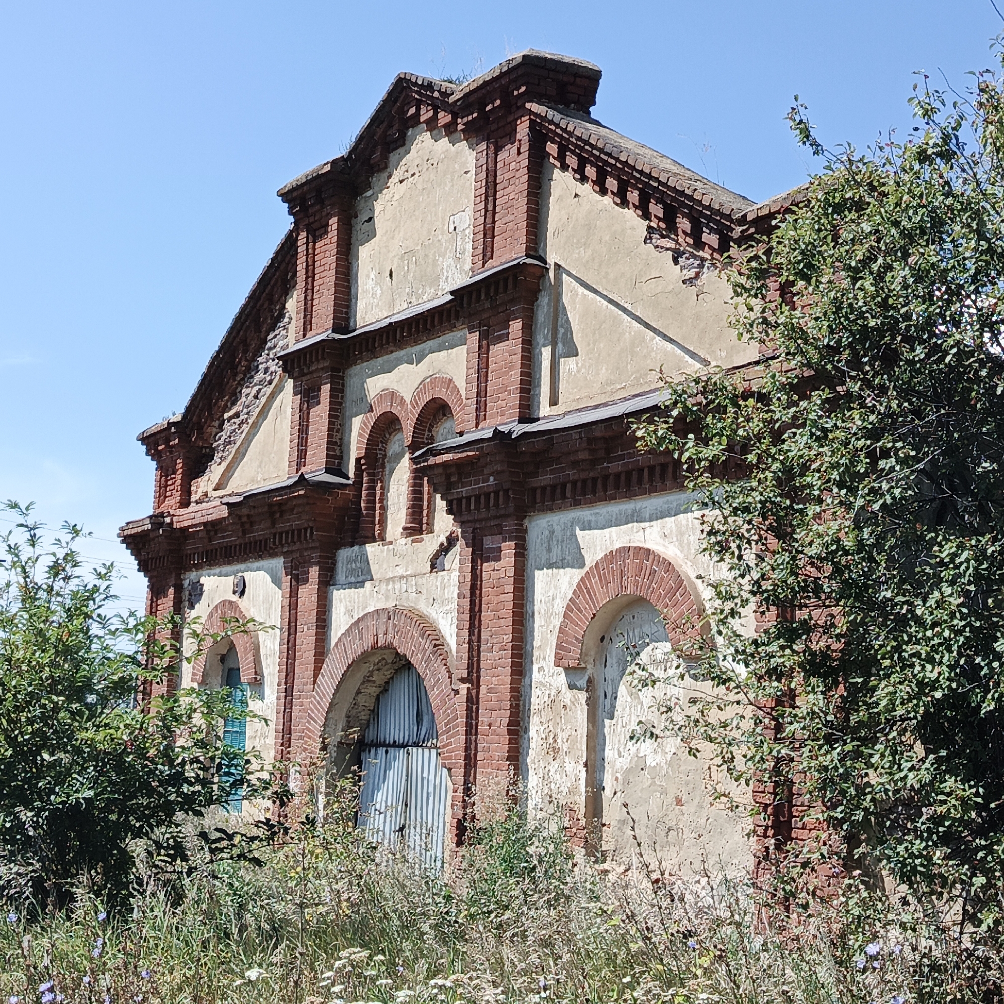 The image depicts an abandoned brick building with a light-colored, weathered facade, overgrown with greenery on the right side. The building features red brick detailing around arched windows and along the roofline, some of which is damaged. Tall grass and various plants surround the structure, with a clear blue sky overhead. The building appears to be a historical industrial or agricultural facility, possibly a former warehouse or factory, located in the village of Tugustemirovo, Russia. There are no people visible in the image.