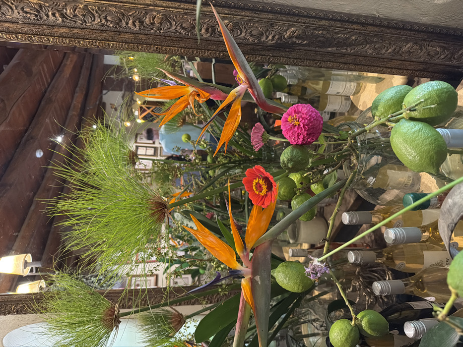 The image depicts an indoor scene within a commercial establishment in Drakenstein Nu, South Africa, characterized by a prominent floral and fruit display in the foreground. Directly in view are several vibrant orange and blue Strelitzia (Bird of Paradise) flowers, numerous clusters of light green papyrus plants (Cyperus), and various smaller red, pink, and purple blossoms, likely Zinnias. Interspersed within these arrangements are bunches of bright green limes on stems, some held in clear glass vases. Below the primary floral arrangement, multiple bottles, predominantly white wine with silver caps, are visible, some directly and others reflected.
A large mirror, framed by an ornate dark wood border on the right, occupies a significant portion of the background. The mirror reflects a segment of the interior, including a person seen from behind, positioned towards the center of the reflection. This individual wears a light blue t-shirt and dark pants. The reflection also shows exposed wooden ceiling beams, suggesting a rustic architectural style, along with overhead lamps emitting a warm glow, and framed items on a wall. A white circular smoke detector is visible on the reflected ceiling. The overall impression is that of a well-lit space, possibly a restaurant, bar, or shop, with a decorative and fresh aesthetic.