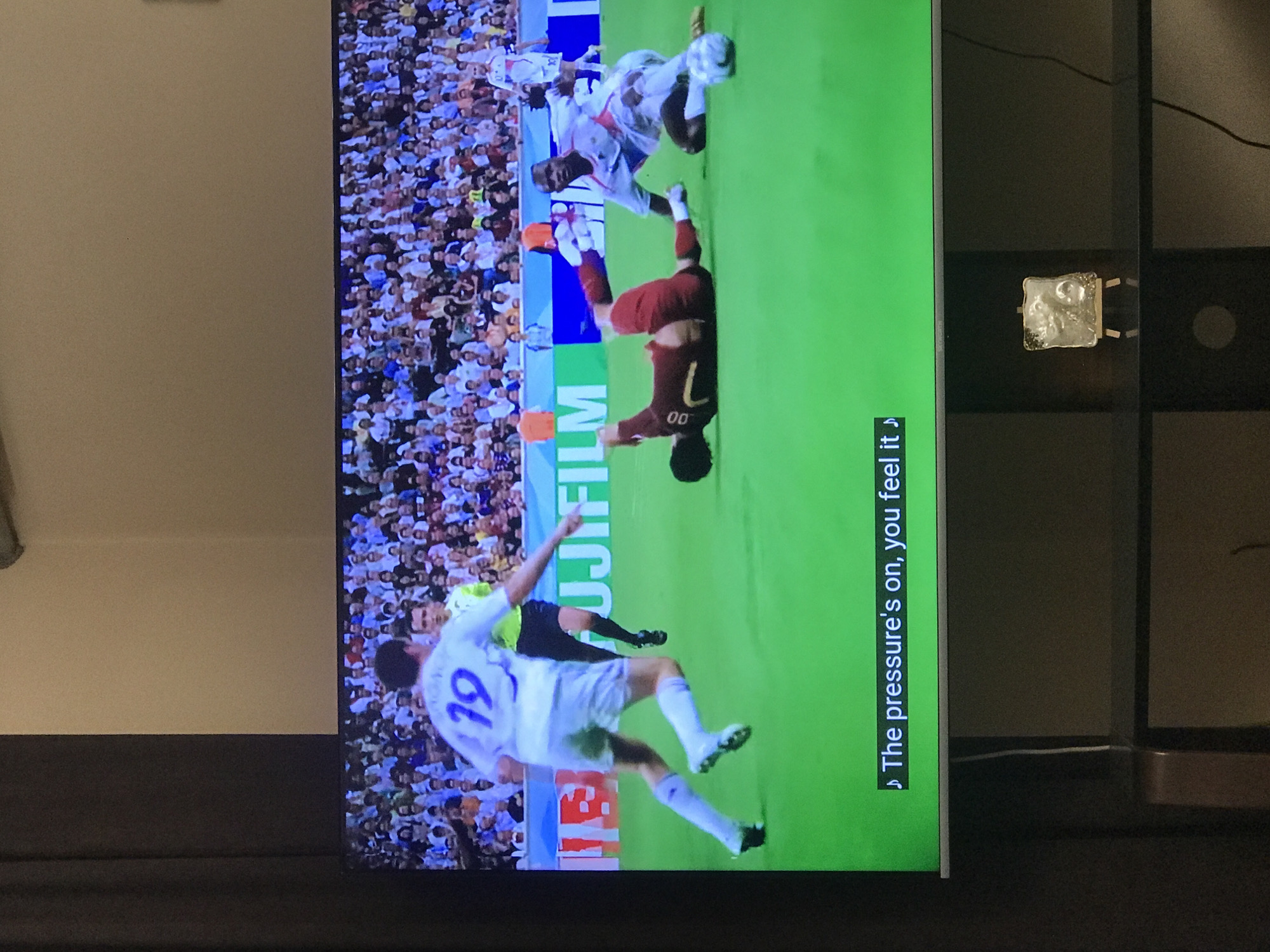 The image shows a television screen displaying a soccer match in progress. A player wearing a white jersey with the number 19 is kicking the ball. Another player wearing a red jersey with the number 00 is falling to the ground. The image is displayed on a television in what appears to be a living room. The room and the soccer game have no known connection to Yerevan, Armenia.
