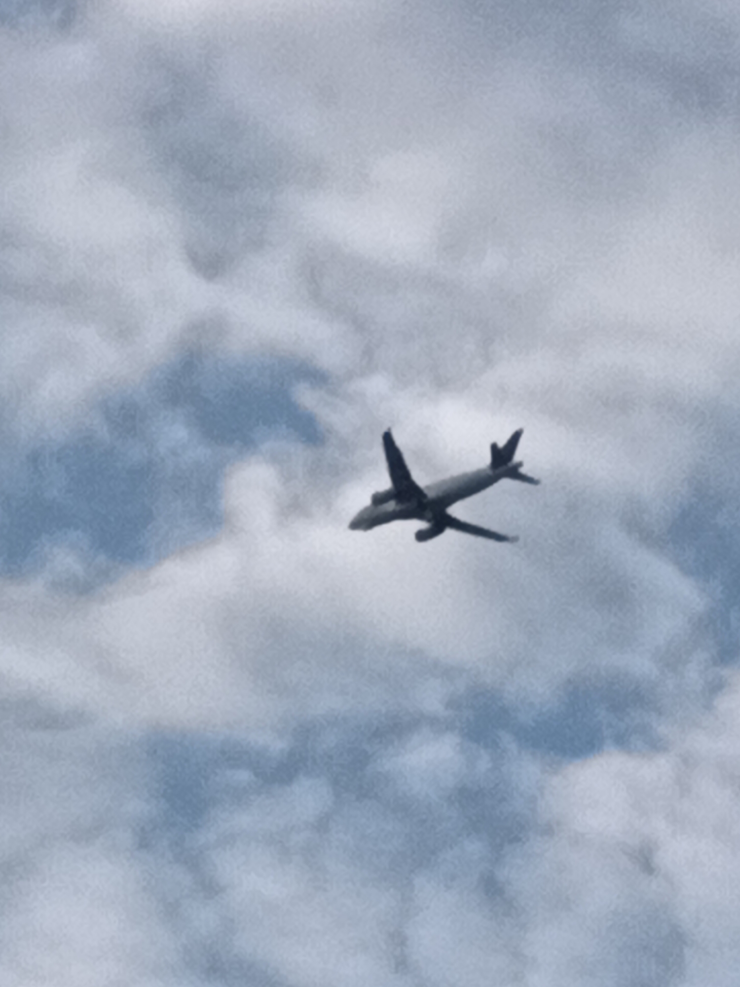 The image shows an airplane flying in the sky, with clouds visible. The airplane is dark in color and appears to be a passenger jet with wings and a tail. The sky is a mix of blue and white, with fluffy clouds obscuring parts of the sky. The lighting suggests it is daytime. The image does not contain any discernible landmarks or features that would indicate its location within the city of Prague, Czechia.
