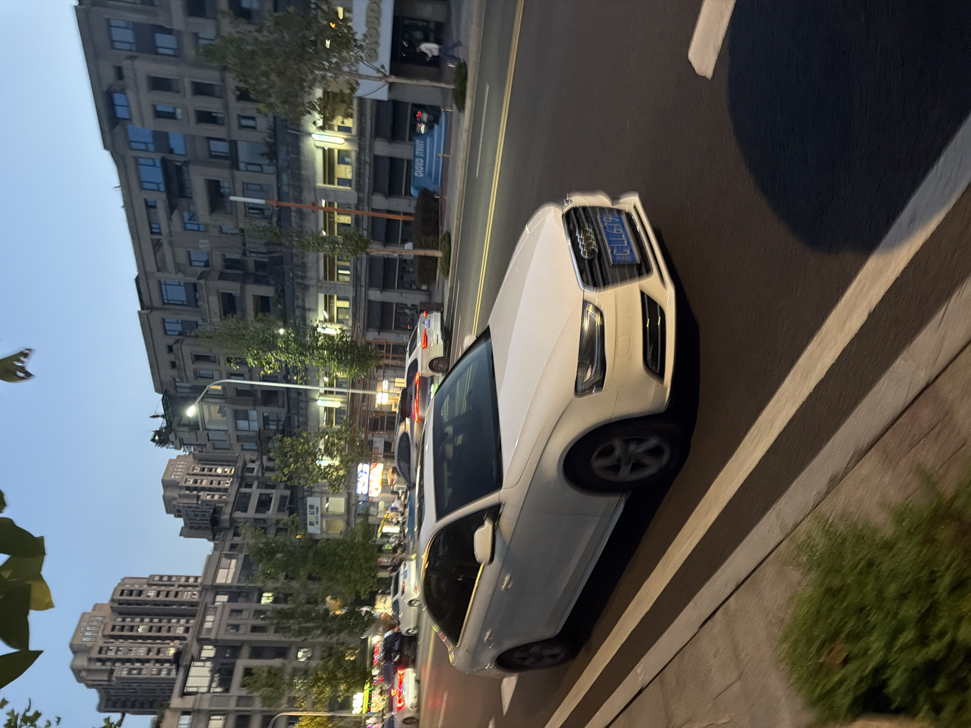A white Audi A4 sedan with license plate C-LL679 is driving on a road at dusk. The car is in the foreground, positioned on the right side of the frame, partially obscuring the road markings and the sidewalk. The license plate is clearly visible and readable. In the background, a street lined with trees and modern buildings can be seen. The buildings are multi-story structures with windows and balconies, suggesting an urban environment. The sky is twilight blue, indicating the time of day. There are other cars visible further down the road, some of which are white sedans. A street lamp is illuminated on the right side of the road. The overall scene depicts a street view in a city, likely Jiangjin, China, given the context.