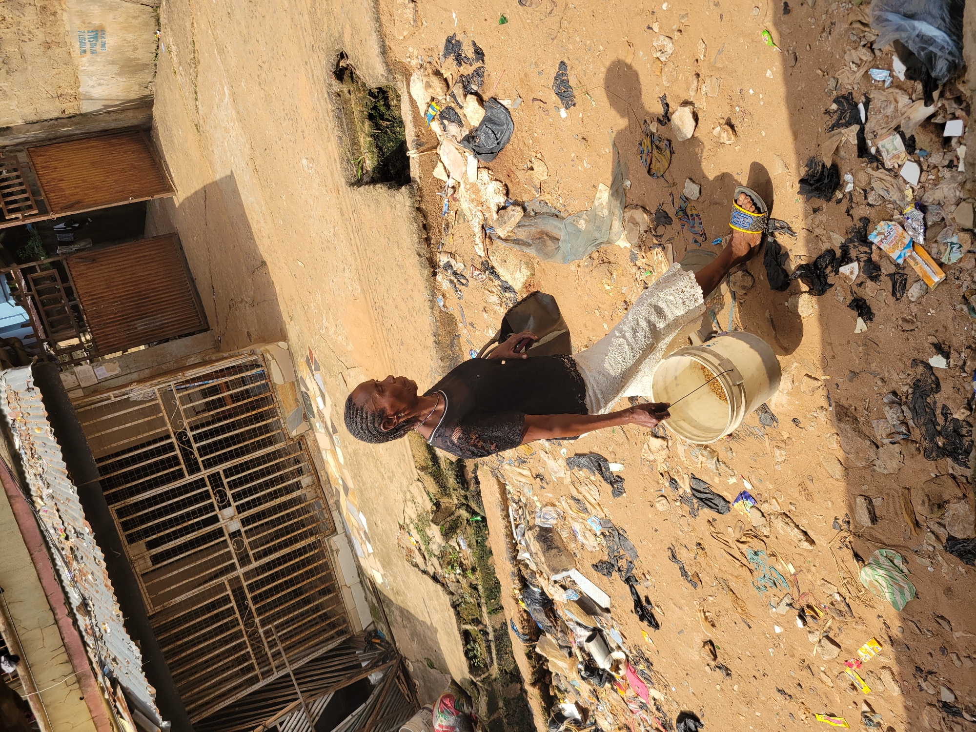 In the city of Unknown, Unknown, an African woman stands in a dirt alleyway littered with trash. The woman is wearing a black lace-trimmed top and a white patterned skirt. She is holding a white bucket in her right hand and a dark purse on her left shoulder. The alleyway is lined with corrugated metal structures and rusty gates. The ground is covered with sand, rocks, and a significant amount of plastic bags, wrappers, and other debris. A drainage ditch with some vegetation is visible in the background. Sunlight casts shadows on the ground.