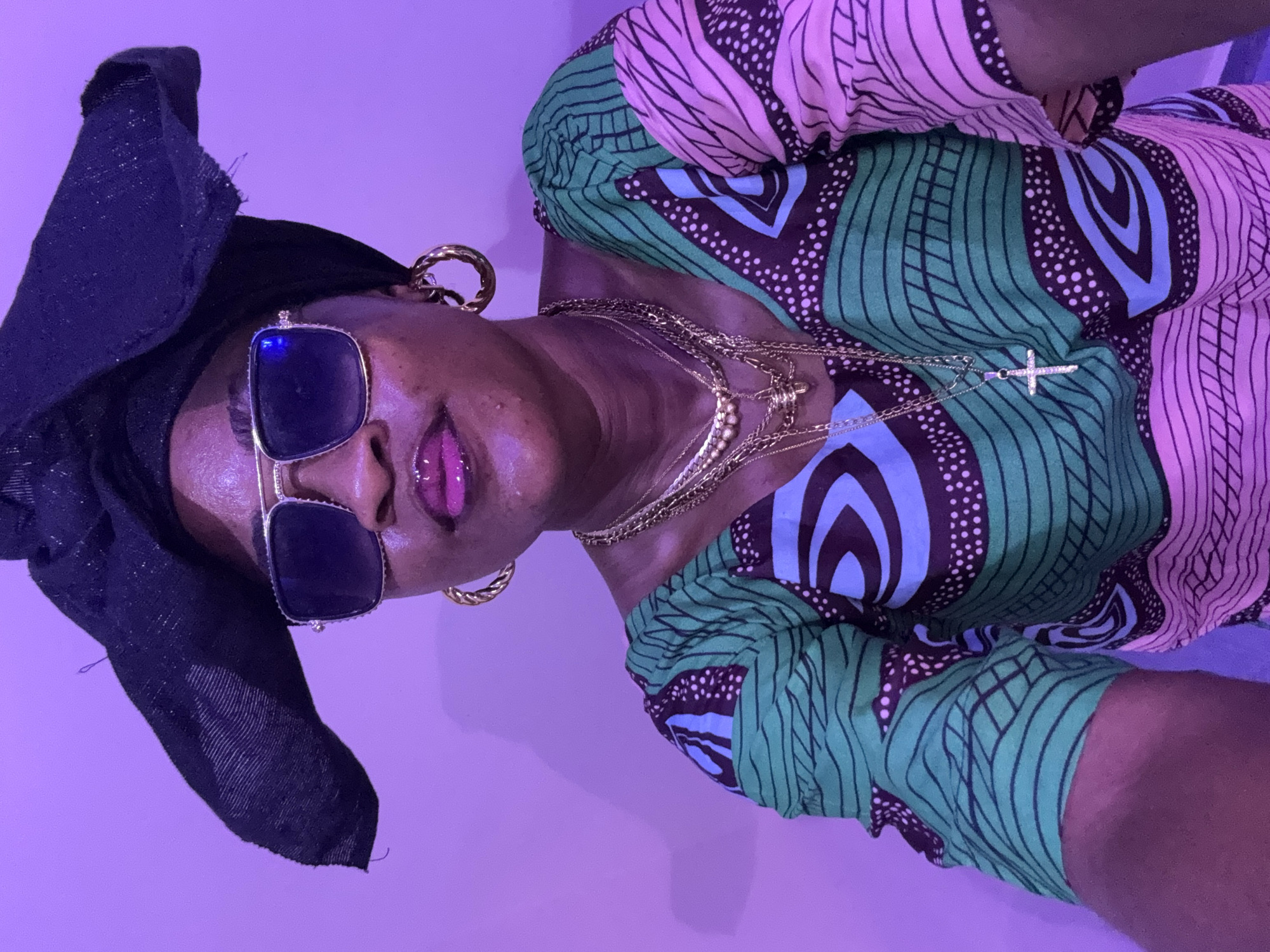 A medium shot captures a Black woman from the shoulders up, seated indoors against a pinkish-purple wall. She wears a black headwrap, gold-rimmed sunglasses, and a colorful garment with green, pink, and blue patterns. Several gold necklaces adorn her neck, including one with a cross pendant. A large gold hoop earring is visible on her right ear. The lighting is dim and casts a purple hue over the scene. No specific details about the location in Ajara, Nigeria, are visible in the image.