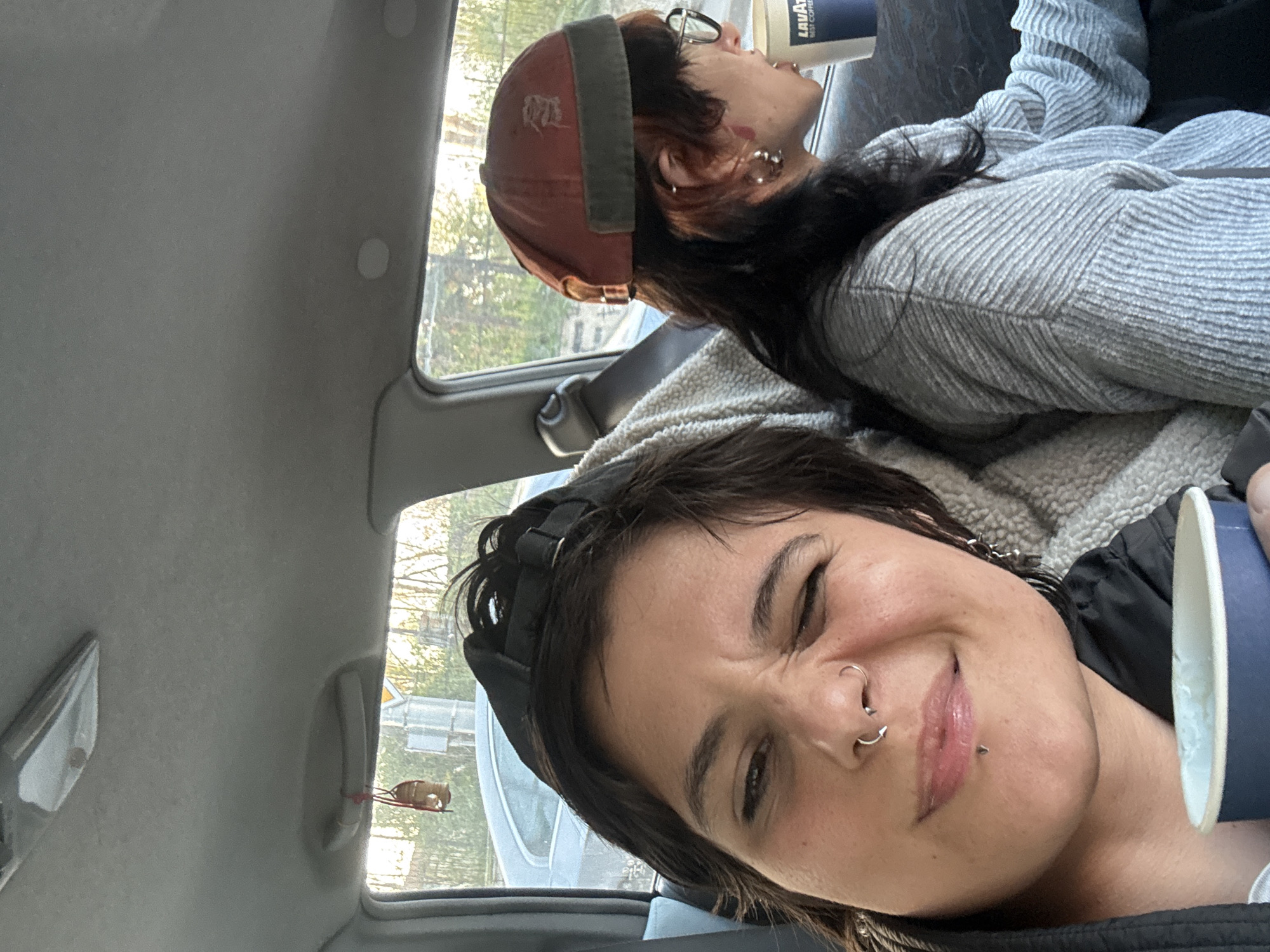 Two individuals are in a car. The foreground person is a woman with short dark hair, wearing a black baseball cap and has multiple facial piercings on the nose and lip, including a nostril piercing and a labret piercing. She is winking. The background person, situated in the passenger seat, has dark hair and is wearing glasses and a red cap. They appear to be drinking from a coffee cup with "Lavazza" printed on it. The environment outside the car window features trees and what appears to be a traffic sign. The car's interior is visible, including the headliner and a small decorative item hanging from the rearview mirror. The location is Sofia, Bulgaria.