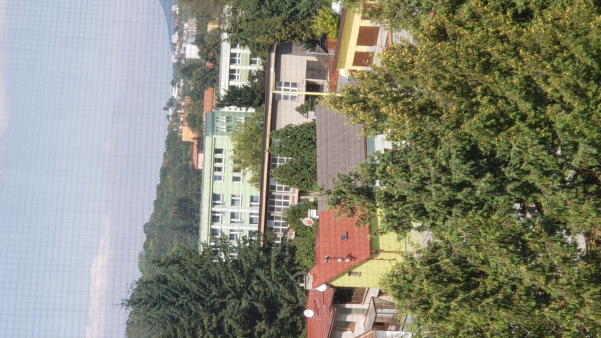 The image depicts an elevated view of a residential area with multiple buildings and dense vegetation, likely taken from a window with a visible mesh screen. In the foreground, several houses with red and brown tiled roofs are visible, interspersed with tall, dark green evergreen trees. Some of these houses have satellite dishes on their roofs. Behind these, a light green, multi-story building with numerous windows is prominent, with a smaller, grey, single-story building with large windows situated in front of it. Further in the background, more buildings of varying heights and colors are scattered across a hilly, tree-covered landscape, suggesting an urban or suburban environment. The sky above is light blue with some white clouds. This scene is consistent with the residential areas found on the outskirts of Prešov, Slovakia, particularly in districts like Sekčov or Sídlisko II, which feature a mix of apartment blocks, family homes, and green spaces.