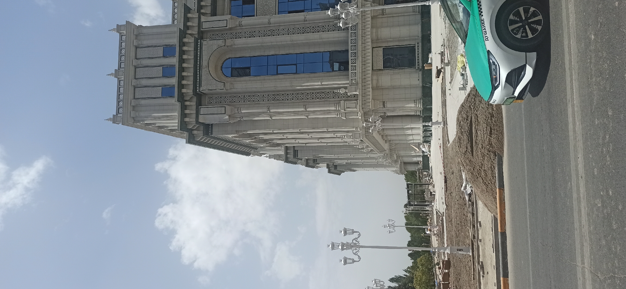 The image shows a street scene in Dushanbe, Tajikistan, with a large, ornate building under construction in the background. The building features classical architectural elements such as columns, arched windows, and decorative stonework. In the foreground, a turquoise and white car, identified as a taxi by Russian script on its side reading "ДУШАНБЕ" (Dushanbe), is parked on the right side of the road. The area in front of the building appears to be a construction site, with piles of sand, paving stones, and scaffolding visible. Several decorative lampposts line the sidewalk. The sky is partly cloudy. No people are clearly visible in the frame.