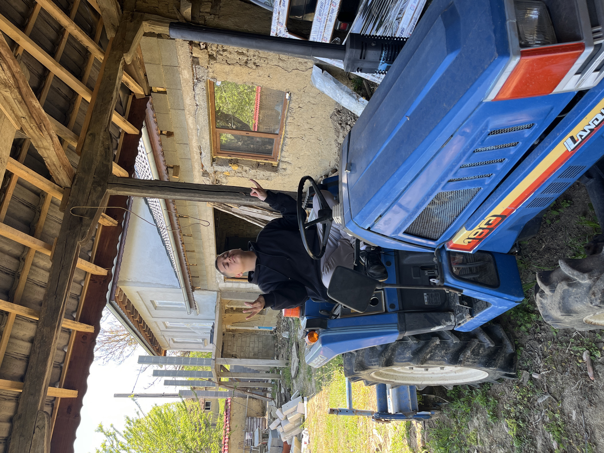 A person is sitting on a blue Landini tractor in what appears to be a rural or semi-rural environment. The tractor is parked on grassy land, near a building under construction. The building is partially exposed and appears to be a two-story structure, with the upper portion freshly plastered or painted. Construction materials are visible near the tractor. The image has elements of an agricultural setting combined with renovation activity. The location is Rish, Bulgaria.