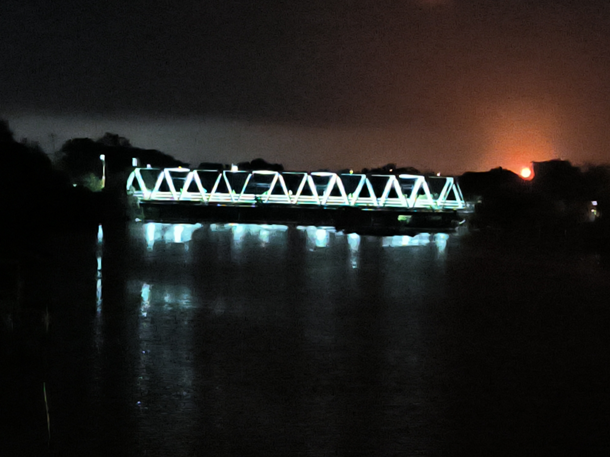 A nighttime view depicts a bridge spanning a body of water, likely a river. The bridge is illuminated with bright, possibly LED, lights accentuating its triangular truss structure. These lights reflect on the water's surface. In the background, a red glow is visible, possibly from the setting sun or city lights, obscured by vegetation. The scene is dark, with limited visibility beyond the bridge and reflections. Based on the location in Lhokseumawe, Indonesia, this is possibly Krueng Cunda Bridge.