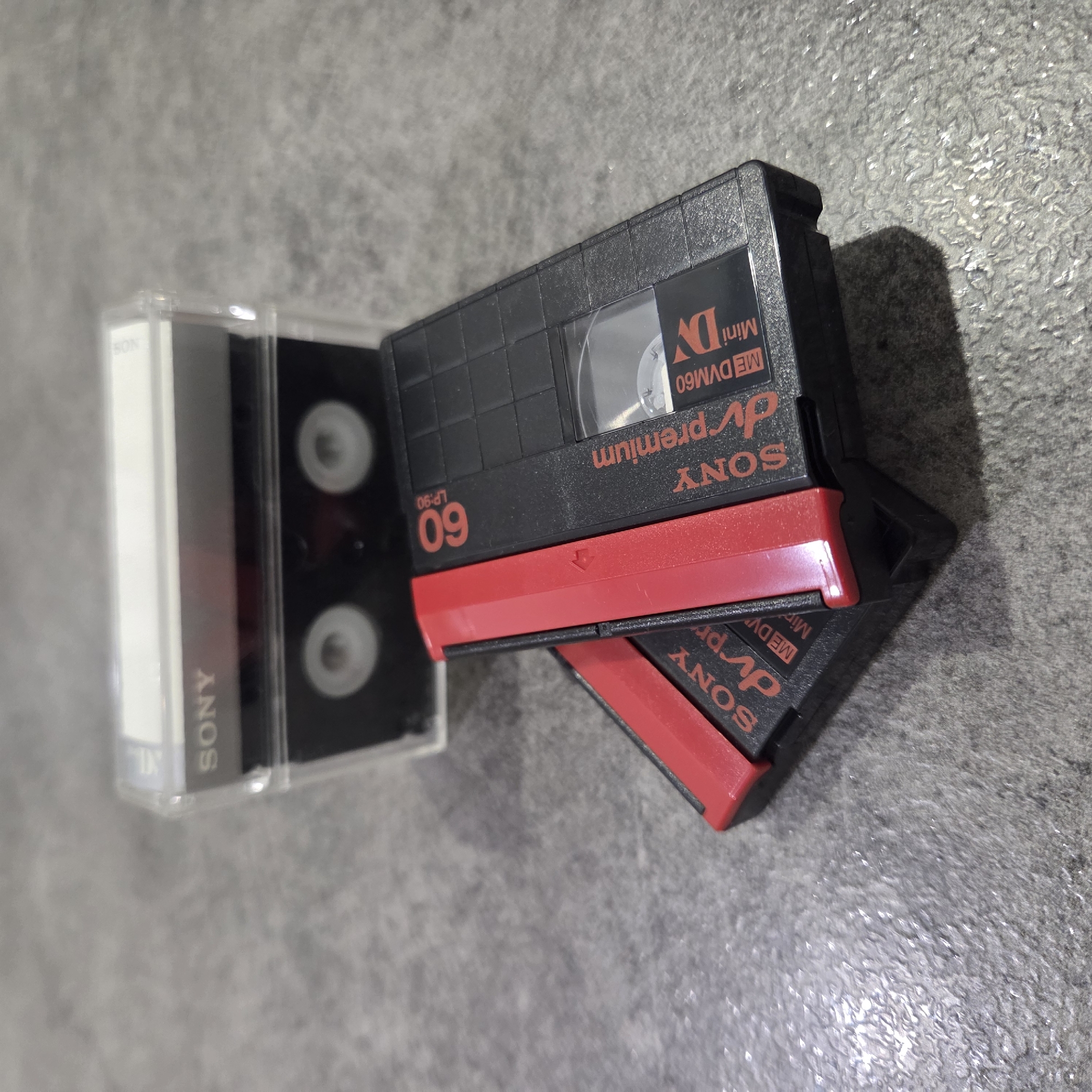 This image features three Sony Mini DV cassettes resting on a textured, grey surface. Two of the cassettes are stacked on top of each other, while a third is enclosed in a clear plastic case and positioned to the left.
The cassettes are primarily black plastic with a characteristic red sliding lock mechanism on one edge. Prominently displayed text on the cassettes includes "SONY," "dv premium," "Mini DV," and "ME DVM60." One cassette clearly shows "60" indicating 60 minutes of recording time, with "LP:90" beneath it, suggesting 90 minutes in Long Play mode. A small arrow symbol is visible on the red lock of the top cassette, likely indicating the opening direction. The clear plastic case also bears the "SONY" brand name on its spine.
The background is a uniform grey, slightly textured surface, possibly a tabletop or floor. Shadows are cast by the cassettes, suggesting directional lighting, likely indoors. There are no people visible, and no specific activity or event is taking place beyond the static arrangement of the objects. The image has a slightly rotated perspective.