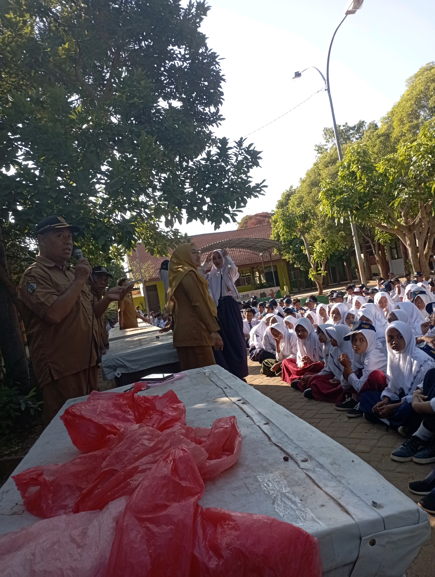 In Banyuurip, Indonesia, a group of individuals, likely students in uniform, are gathered outdoors for an event. The scene is set under a clear sky and surrounded by trees. A man in a brown uniform with a cap is speaking into a microphone, addressing the audience. Beside him, another man in a similar uniform gestures with his hand. A woman in a light brown hijab and uniform stands to the right, facing the seated crowd. The students, predominantly wearing white hijabs and uniforms with red skirts, are seated on the ground, attentively listening. Some students are looking towards the speakers, while others appear to be engaged in conversation amongst themselves. In the foreground, a white table is covered with red plastic bags, obscuring much of its surface. The overall atmosphere suggests an informal outdoor gathering or assembly, possibly at a school or community event.