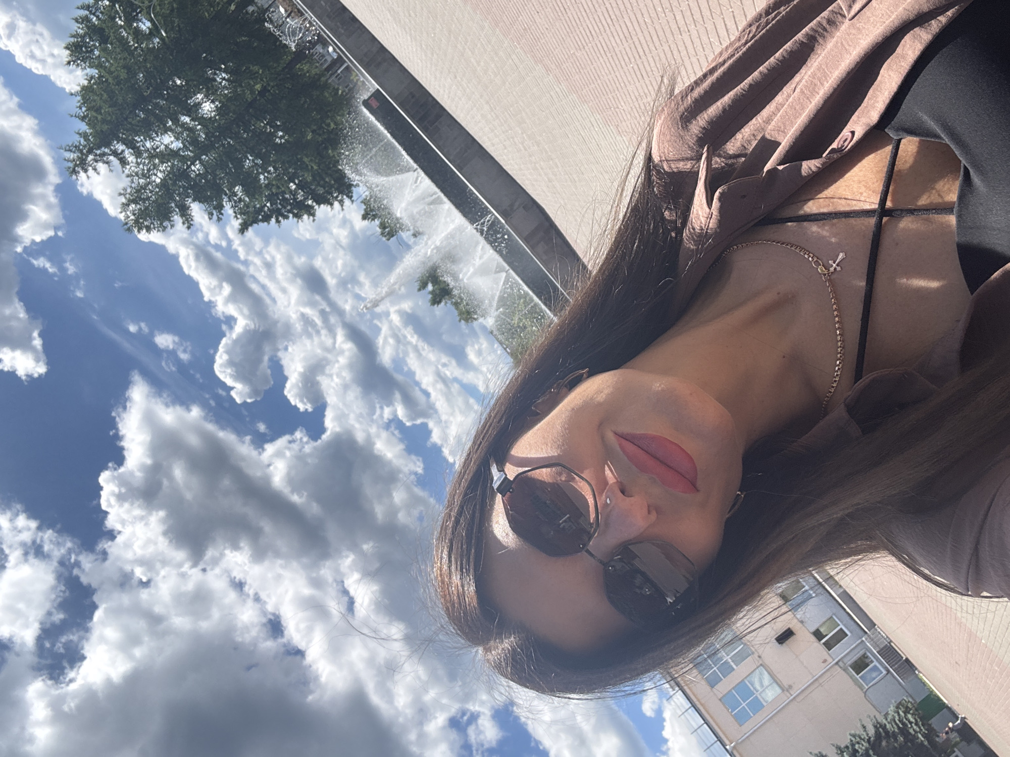 A woman with dark hair and sunglasses is taking a selfie in front of a fountain in Chaikovsky, Russia. She is wearing a brown shirt and a black top. The fountain is in the background, with trees and buildings visible on either side. The sky is blue with white clouds.