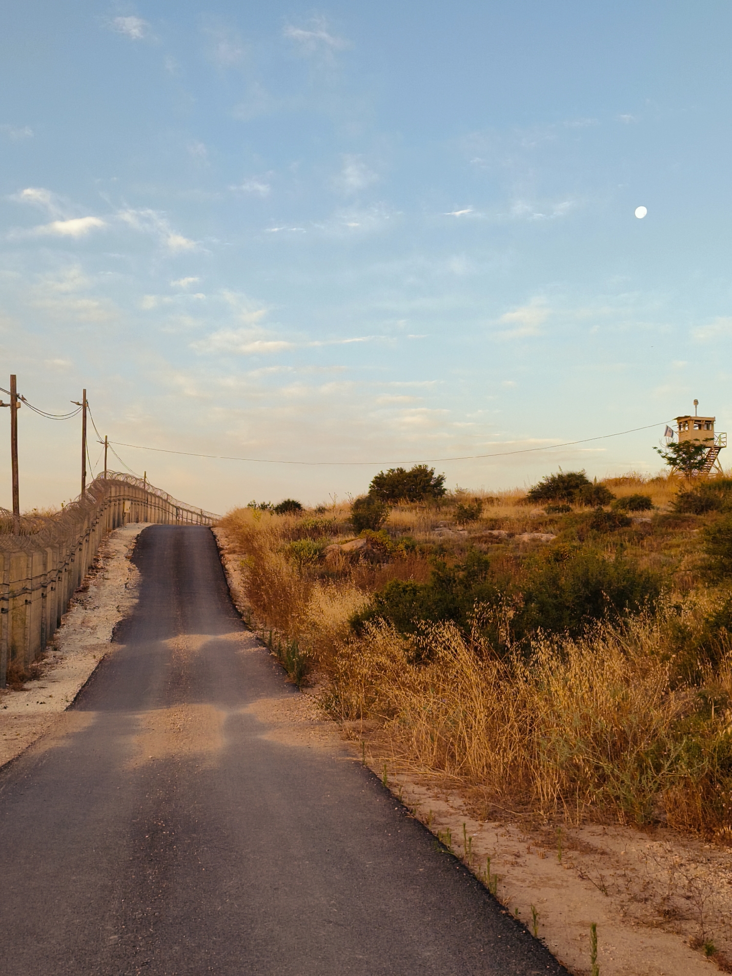 This image depicts a rural landscape under a twilight sky, featuring a road, a security fence, and a watchtower. A narrow, dark gray paved road ascends a gentle slope, curving slightly uphill into the distance. To the left of the road, a tall concrete and barbed wire security fence runs parallel to the road, also climbing the incline. Wooden utility poles with power lines follow the fence line.
The surrounding terrain is covered with dry, golden-brown grasses and scrub brush, characteristic of an arid or semi-arid environment. Scattered green bushes and small trees are visible among the dry vegetation, particularly on the right side of the road. Further up the hill on the right, a wooden watchtower stands tall, featuring an enclosed cabin at its top and an external ladder or staircase. A flag with distinct blue, white, and red vertical stripes, identifying it as the French flag, flies atop the watchtower.
The sky is a clear, light blue with scattered wispy white clouds. A bright, full or nearly full white moon is visible in the upper right portion of the sky, indicating either dawn or dusk. The warm, golden light illuminating the landscape suggests the "golden hour" of late afternoon or early evening. No human activity or events are taking place; the scene conveys a sense of quiet solitude. The presence of the security fence and watchtower, along with the French flag, suggests a controlled area, perhaps a border, military post, or peacekeeping zone, located within the Lachish Regional Council in Israel.