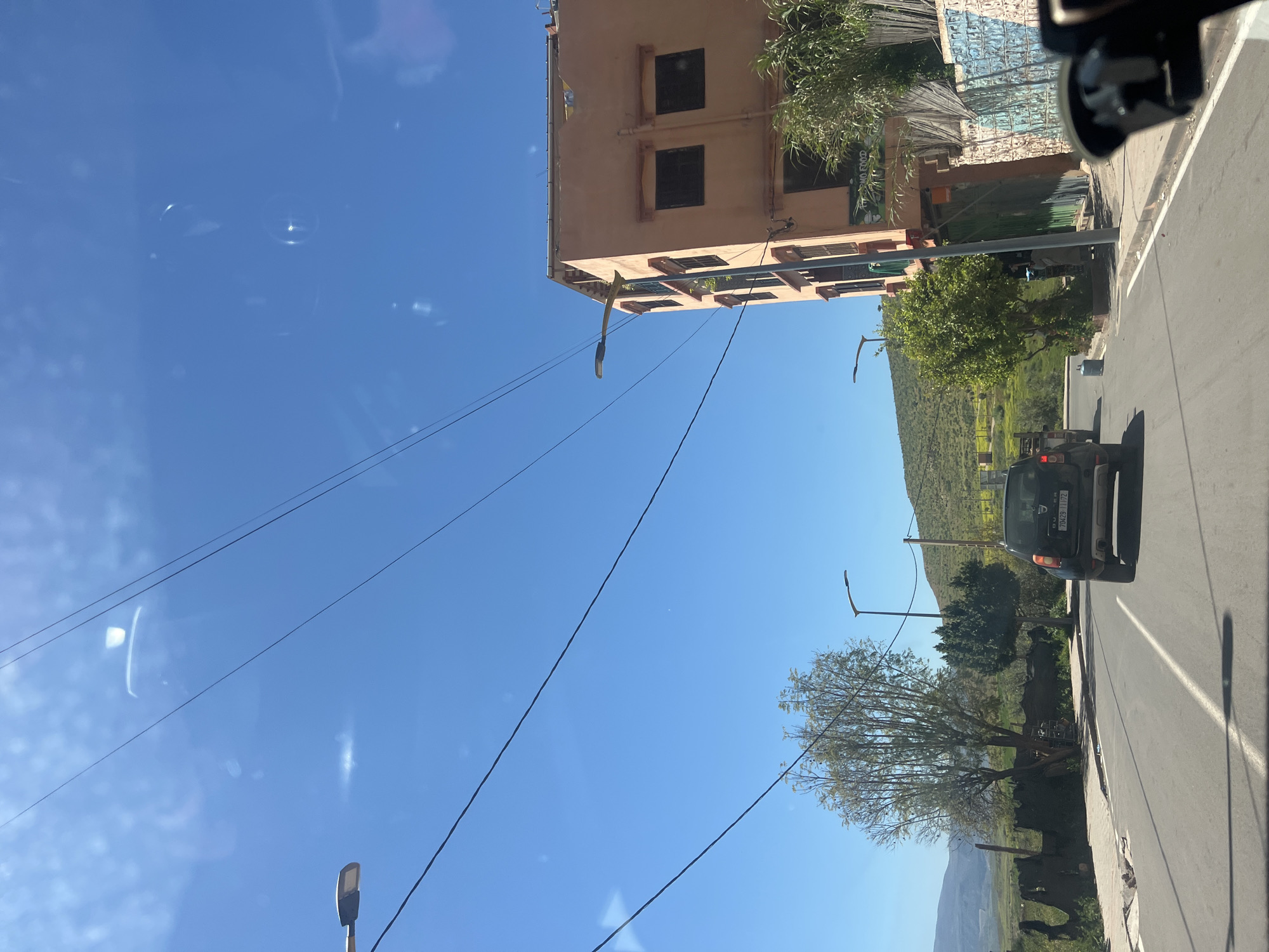 The image shows a street in Ouaouizert, Morocco, viewed from a vehicle. A two-story building with multiple windows is on the left side of the frame. Power lines stretch across the clear blue sky. A black car drives on the right side of the road, which is lined with trees and vegetation. A hill is visible in the background. Street lights are positioned along the road.
