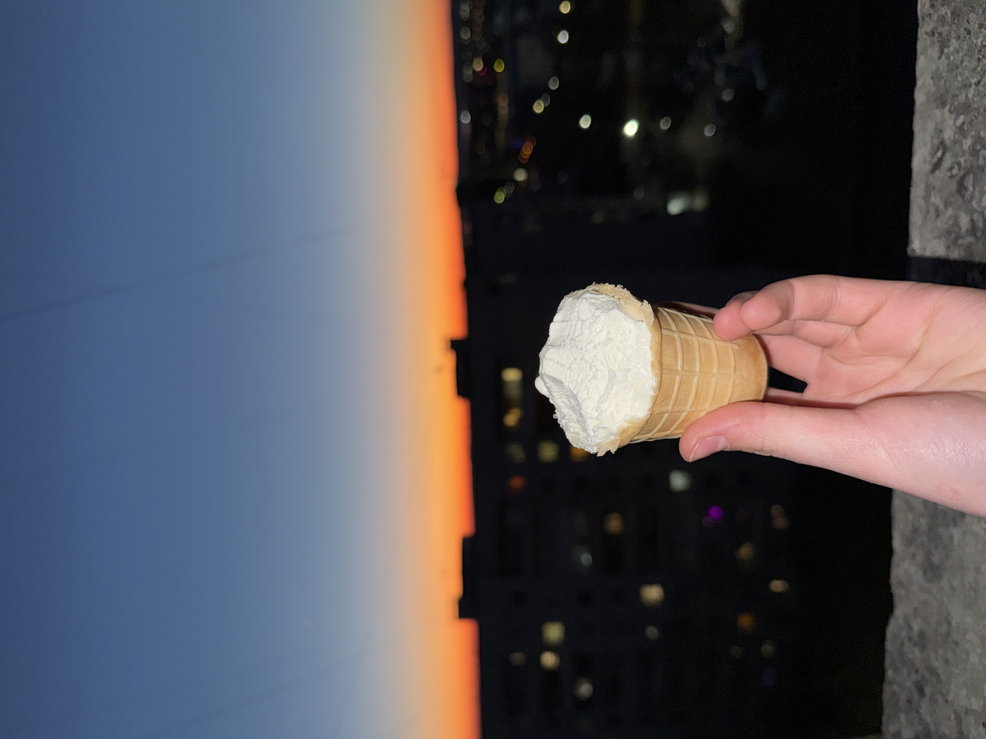 The image presents a hand holding an ice cream cone against a cityscape backdrop at dusk. The sky transitions from a light blue at the top to a vibrant orange near the horizon. Buildings are silhouetted against the fading light. The ice cream cone is filled with a white substance, presumably ice cream or a similar frozen dessert. The hand holding the cone appears to be fair-skinned. Small, blurred lights suggest an urban environment. The location could be the city of Chaikovsky, Russia.