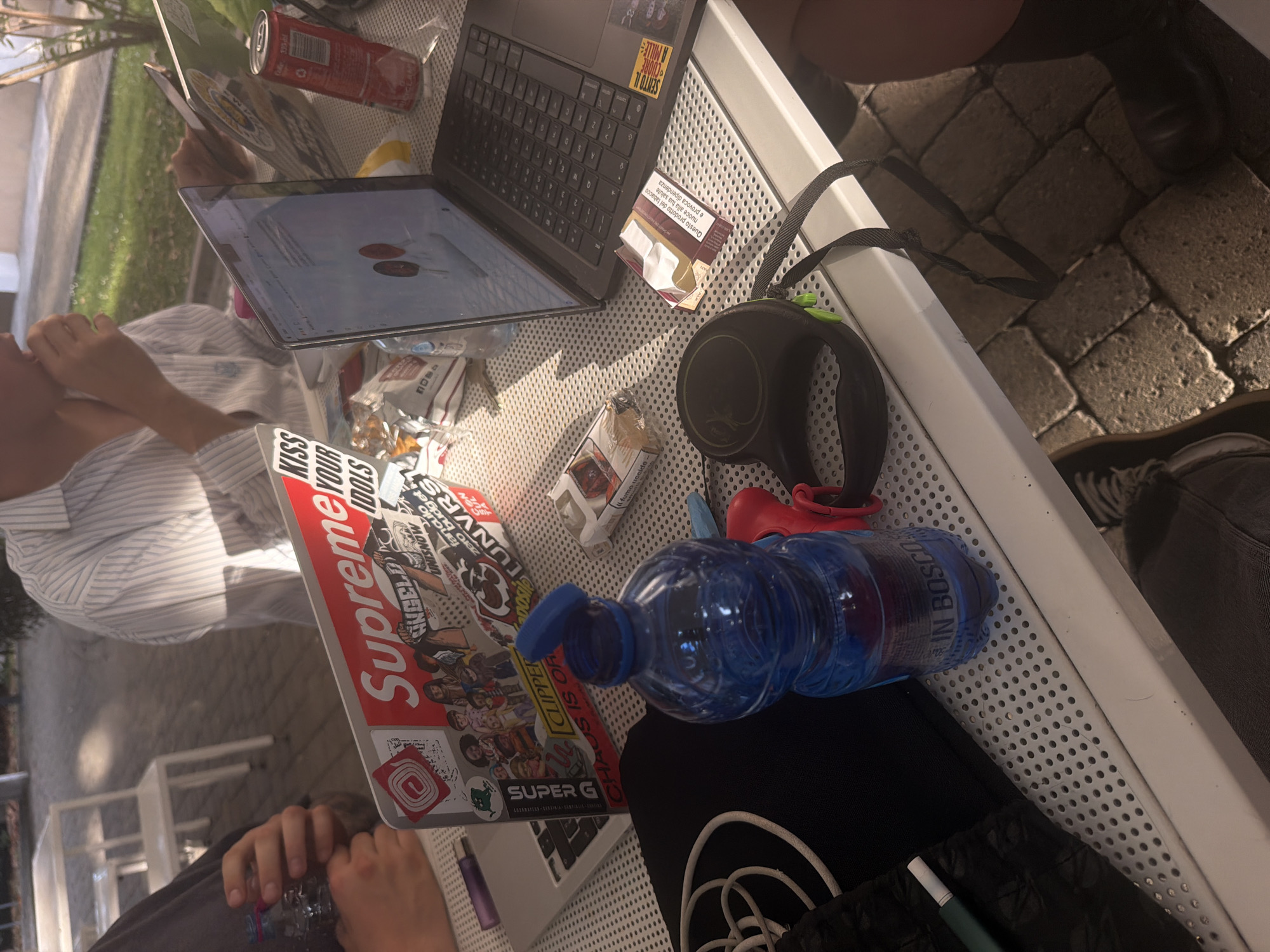The image presents an outdoor scene, likely a cafe or an al fresco study spot in Milan, Italy, as indicated by Italian text on various items. Two individuals are present, engaged with laptops on a white, perforated metal table during a bright daytime, possibly afternoon, setting.
One person, wearing a light-colored striped shirt, is visible from the chest up, with their hand resting near their face, appearing focused on a laptop screen displaying a web page. The hands of a second person are visible at the bottom of the frame, holding a blue plastic water bottle.
The table is laden with personal items. Two laptops are open; one is adorned with numerous stickers including "Supreme," "KISS YOUR IDOLS," "CLIPPER," "SUPER G," "ANGELD," "zero budget," "fanculo," "UNISNOW," "SENTO IL CUORE A MILLE," and "LADOSE CUNVRS." A red 330ml Coca-Cola can sits near one laptop. A partially open pack of cigarettes is visible, bearing the Italian health warnings "Questo prodotto del tabacco nuoce alla tua salute e provoca dipendenza" (This tobacco product harms your health and causes addiction) and "fumo uccide" (smoking kills). A blue plastic water bottle, labeled "IN BOSCO," is also on the table, next to a black retractable dog leash and a small red item. The ground beneath the table is covered with paving stones, and greenery is visible in the background, reinforcing the outdoor environment. The scene suggests a casual and focused outdoor work or study session.