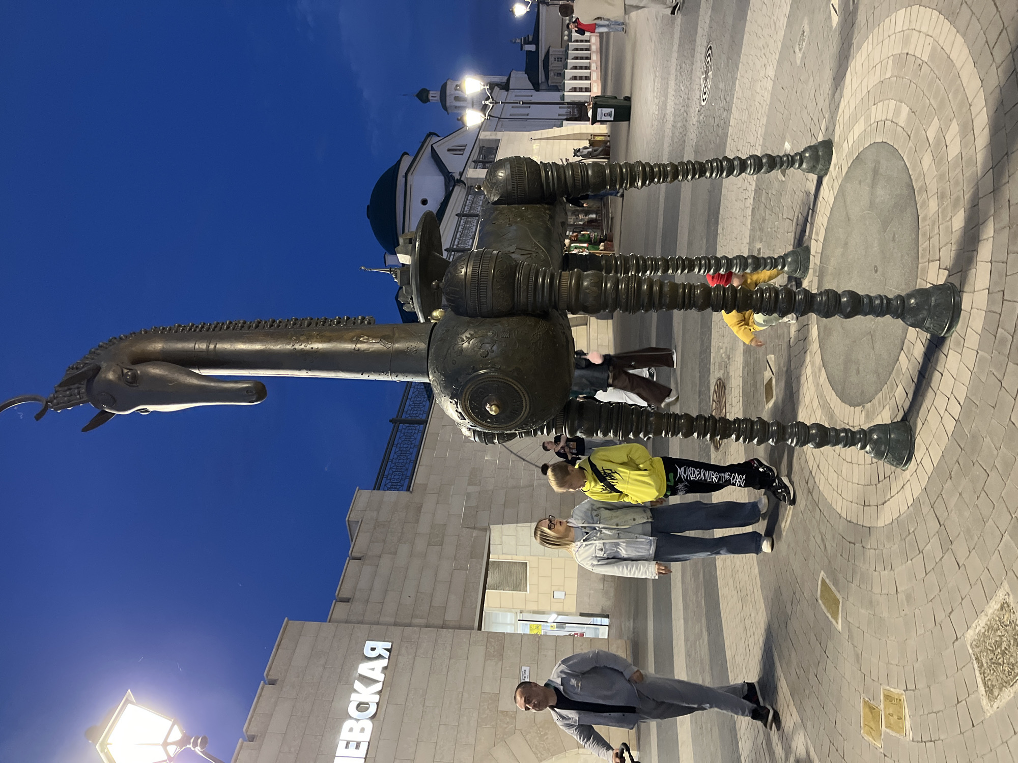 This image captures an outdoor scene at dusk in Kazan, Russia. The central subject is a large, intricate bronze sculpture of a horse with mechanical-looking legs. The sculpture's design is detailed, featuring segmented legs and a stylized horse head.
In the foreground, several people are visible, seemingly interacting with or observing the sculpture. A man in a gray suit walks past, looking down. Behind him, two women stand near the sculpture. One woman wears a light-colored jacket and jeans, while the other is dressed in a bright yellow hoodie and dark pants. Further in the background, other individuals can be seen, including a person in a red jacket taking a photo.
The setting appears to be a public square or pedestrian area, paved with intricate stone patterns. Behind the sculpture and people, a modern building with a light-colored facade is visible. The building has large, illuminated letters that spell "НЕВСКАЯ" in Russian. Overhead, streetlights illuminate the area, suggesting it is evening. The sky is a deep blue, indicating twilight. The overall atmosphere seems calm and urban.