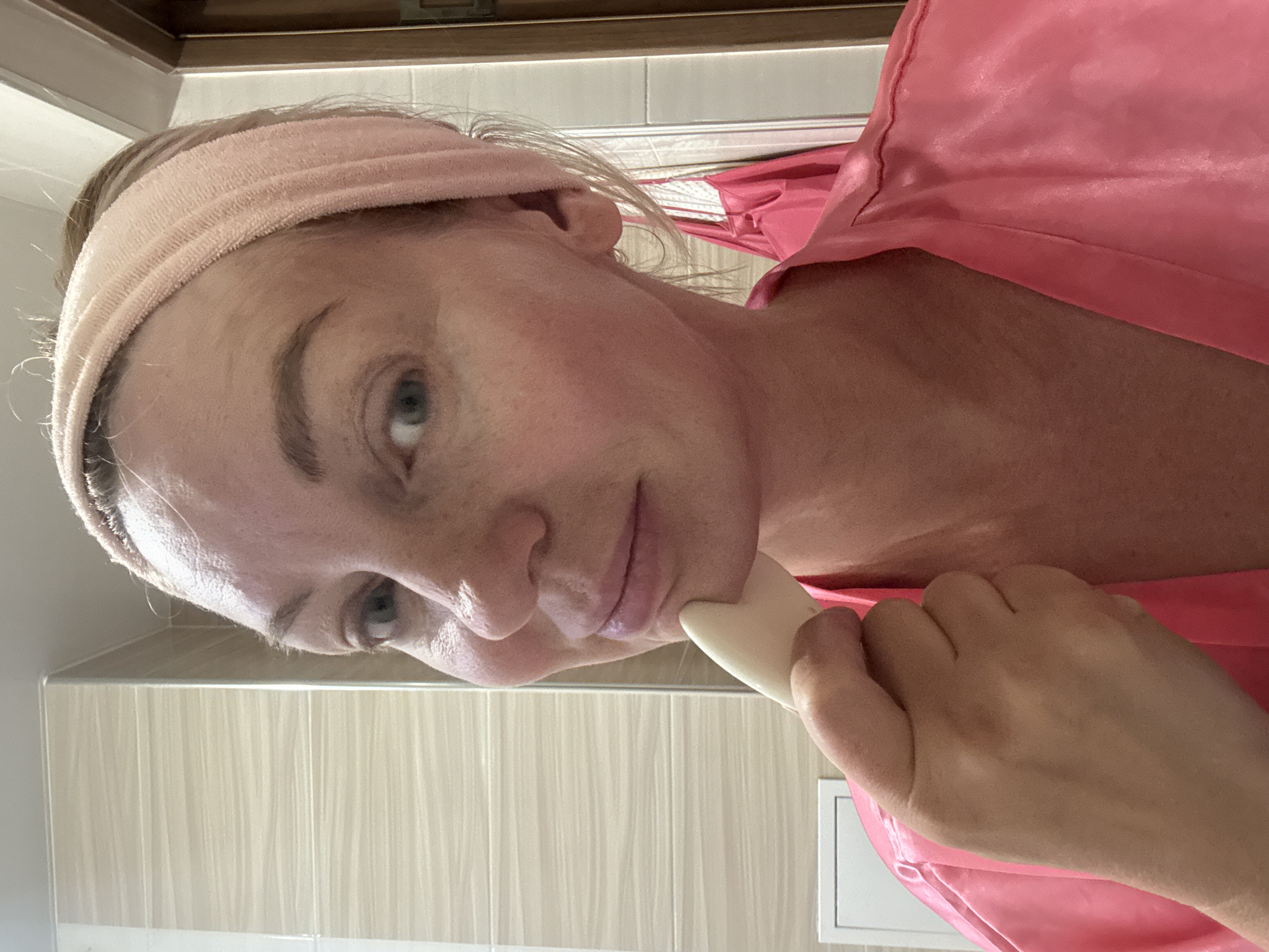 A woman is shown from the chest up, looking directly into the camera, in what appears to be a bathroom. She is wearing a pink satin robe and a pink terrycloth headband. Her right hand is raised to her chin, holding a light-colored, stone-like facial gua sha tool. She has fair skin, light brown hair, and blue eyes. Her makeup appears natural, with blush visible on her cheeks. The background shows tiled walls, suggesting a bathroom setting. The image does not contain any information that could place it in Sochi, Russia.