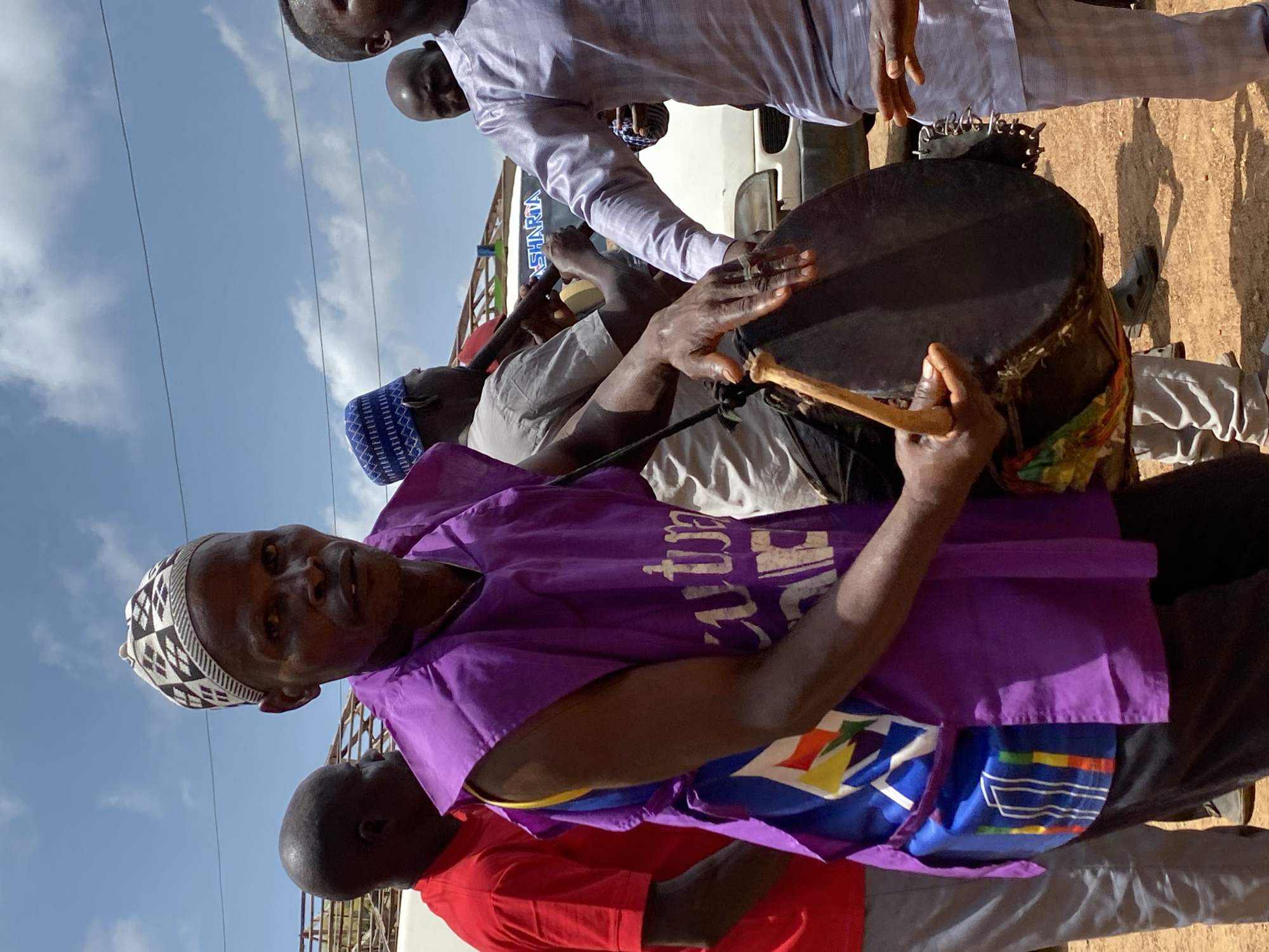 In Maigizo, Nigeria, a group of people are gathered near a white vehicle under a partly cloudy sky. The main subject is an African man wearing a purple vest and a patterned head covering. He is holding a drum and a stick, appearing to play it. Other people, including one in a blue patterned hat, are nearby. The location appears to be outdoors, possibly on a roadside.