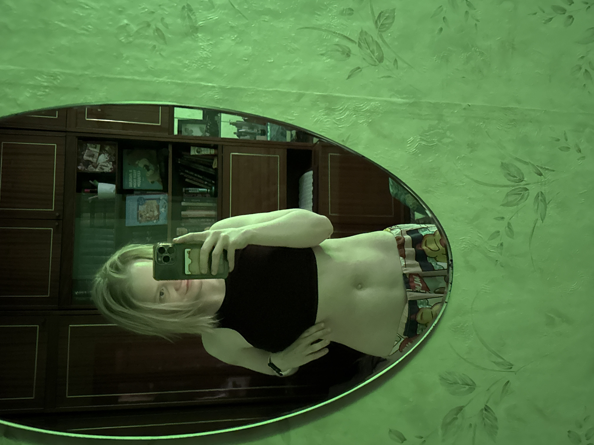 A mirror reflects a person holding a phone, dressed in a black top and patterned shorts, with a bookshelf visible in the background. The lighting casts a green hue over the scene. The location cannot be determined from the image.