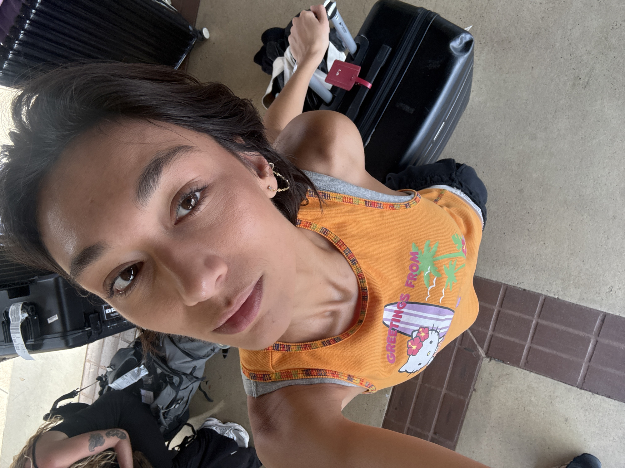 A high-angle selfie captures a woman looking directly into the camera, positioned within a transit area in Lihue, United States of America.
The primary subject, a woman with dark brown hair and tanned skin, occupies the central and lower-right portion of the frame. She wears an orange tank top with grey trim and a multi-colored pattern along the neckline and armholes. The tank top features a graphic depicting Hello Kitty holding a surfboard, with palm trees and seagulls in the background, accompanied by the text "GREETINGS FROM". She exhibits small gold-colored hoop earrings in both ears, with an additional smaller stud in her right ear. Her expression is neutral, and her gaze is directed towards the camera.
In the upper-left background, a second individual is partially visible, seated or crouched. This person has blonde or light brown curly hair and is wearing a black top. Tattoos, including one resembling a creature or tribal design on the upper right arm and another smaller tattoo below it, are visible. A foot wearing a grey and white athletic shoe is also visible.
The ground consists of light beige or concrete-like tiles, traversed by a band of dark reddish-brown rectangular tiles arranged in a pattern. Multiple pieces of luggage are present. A black hard-shell rolling suitcase with a red luggage tag is positioned to the right of the main woman. In the background, several grey and black backpacks or duffel bags are on the floor near the second individual. A large black ribbed suitcase is visible in the upper-right background, and another black piece of luggage is in the upper-left background. The scene is brightly and evenly lit, indicative of an indoor or covered outdoor transit environment.