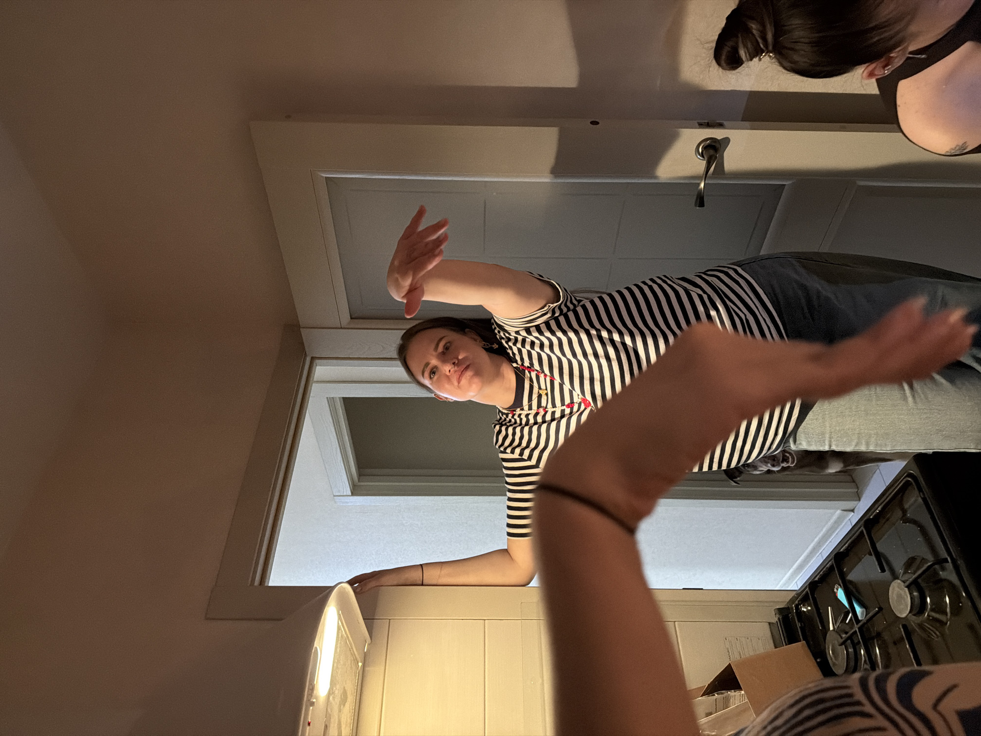 A low-angle shot looking up at two women in what appears to be an indoor kitchen environment. One woman is wearing a black and white striped shirt and jeans and is positioned in front of a doorway or archway. She has one arm extended towards the top of the frame. A second woman is to the right of the frame and partially visible. She has dark hair pulled into a bun. Parts of a stove, countertop, and cabinets are visible at the bottom of the frame. The location is not discernible from the image.