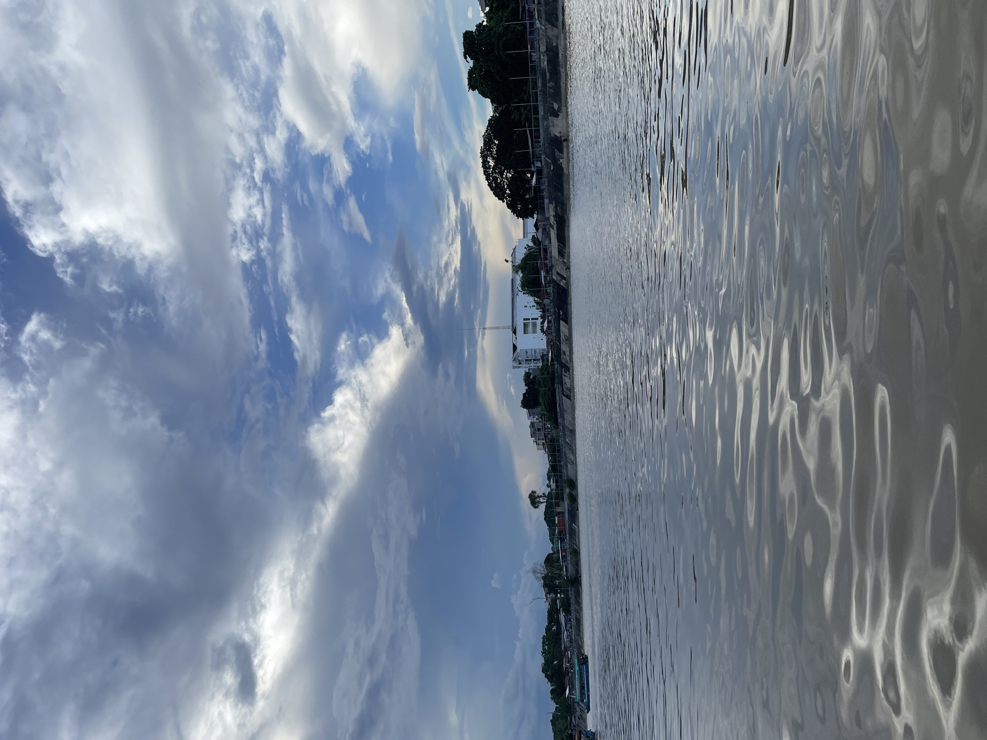 The image depicts a serene river scene in Davao City, Philippines. The murky water reflects the dramatic, cloud-filled sky, with a mix of dark storm clouds and brighter, sunlit patches. Along the riverbank, a line of buildings and trees forms the cityscape. A few boats are visible on the water, one of which is a small passenger boat with a turquoise canopy. The overall impression is one of a quiet, overcast day by the river.