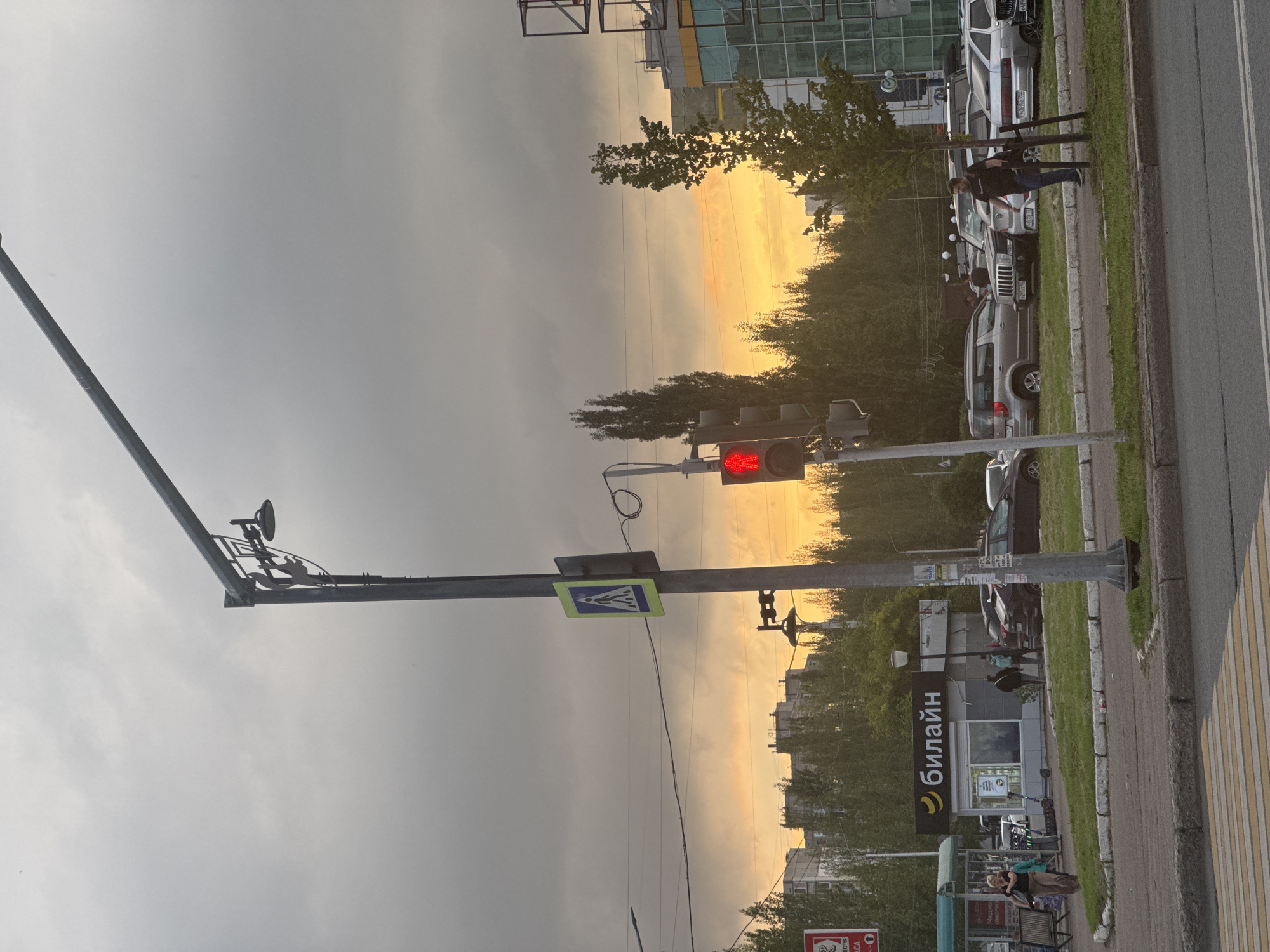 The image depicts an outdoor scene at dusk in Ufa, Russia. A traffic light is red, indicating a stop. A pedestrian crossing sign is visible. Several cars are parked in the background, and some people are walking. A street lamp stands prominently in the foreground. A commercial building with a glass facade is located to the right. The sky is overcast with hues of orange and yellow from the setting sun. The overall scene suggests a typical urban environment with signs of ongoing activity despite the fading light.