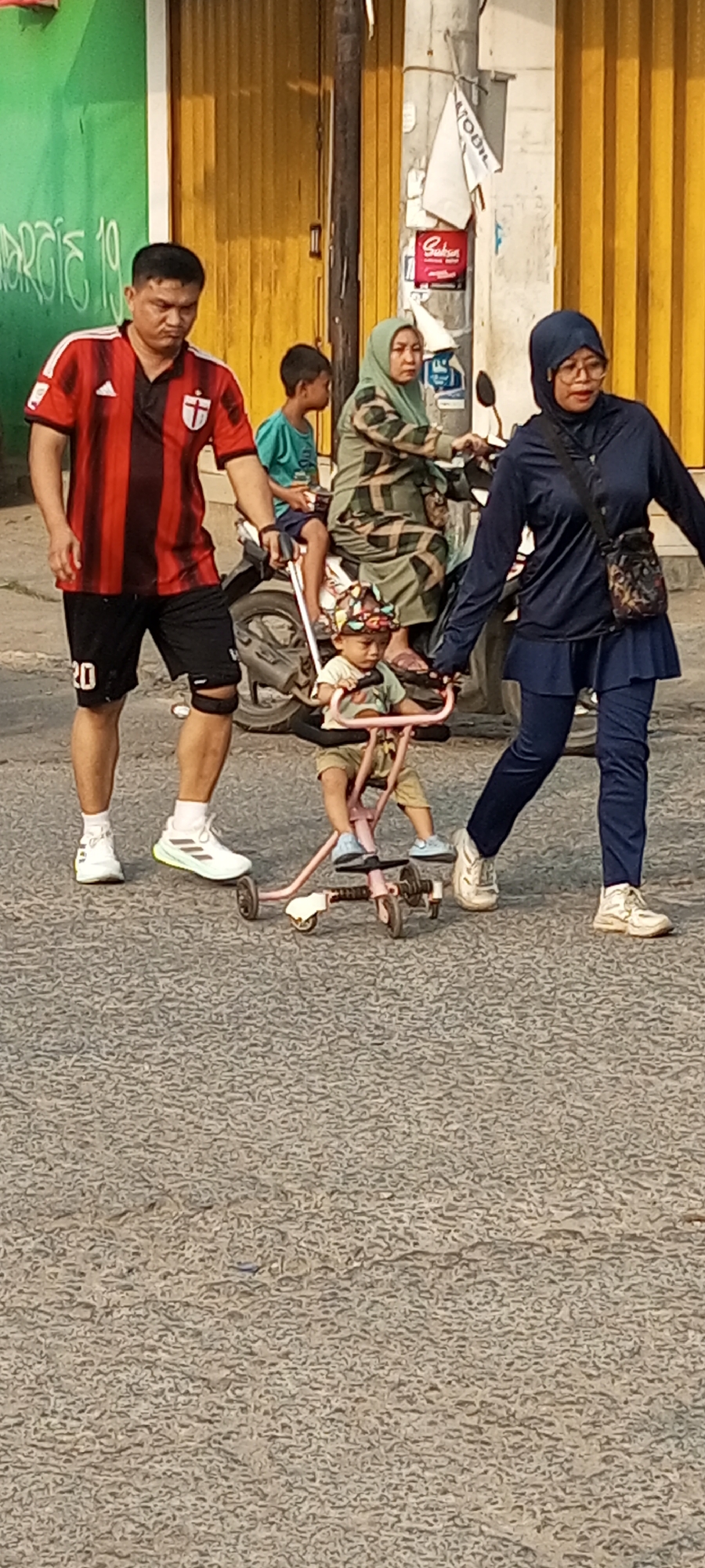 In the city of Setu, Indonesia, a group of people are walking on a paved street. A man wearing a red and black striped shirt and black shorts walks on the left side of the frame, with the number "20" visible on his left leg. Next to him, a young child sits in a pink stroller. Behind the man and stroller, a woman in a camouflage-patterned hijab rides a motorcycle, with another young child sitting behind her. To the right of the woman on the motorcycle, another woman wearing a dark blue hijab and matching long-sleeved shirt and skirt walks, holding the stroller's handle. The background features a yellow corrugated metal wall and a green wall with white lettering. The pavement has a textured, asphalt-like surface.