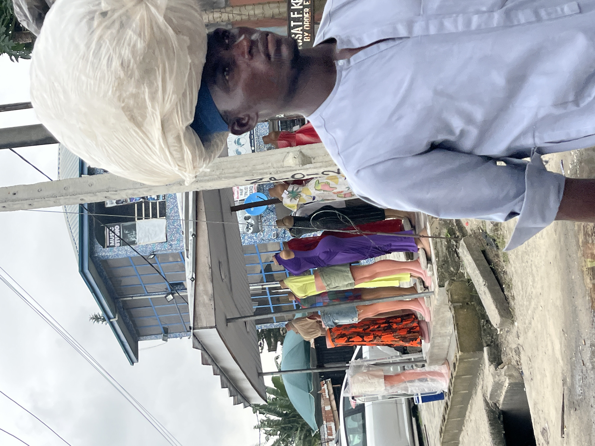 A man in Ado, Nigeria, carries a large, white plastic bag of goods on his head. He is wearing a light blue traditional garment with a mandarin collar. He stands in the foreground, facing right, with his head slightly bowed. Behind him, a row of mannequins displays colorful clothing, including a bright orange patterned skirt, a yellow top, and a purple dress. A green umbrella is visible behind the mannequins, and a white car is parked to the left. A building with blue accents and the sign "MUSIC STUDIO" is in the background, with power lines crisscrossing the sky. The scene appears to be a street market or commercial area.