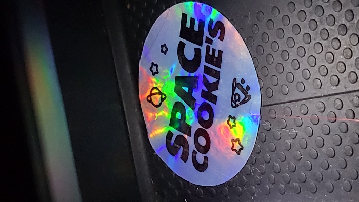 A circular holographic sticker is positioned on a dark, textured surface. The sticker features the black uppercase text "SPACE COOKIE'S" and outlines of celestial objects: a bell-shaped planetoid, multiple stars, and a rocket ship. Iridescent rainbow light reflects across the sticker's surface. The underlying surface is dark grey or black, characterized by a repeating pattern of raised circular nubs, consistent with an anti-slip mat or flooring. A subtle linear indentation or mark runs vertically through the lower half of the sticker. Fine, light-colored fibrous debris is visible on the textured surface adjacent to the sticker. The upper portion of the image is dark, exhibiting a distinct horizontal band of diffused rainbow light. This scene is located in Magaluf, Spain.