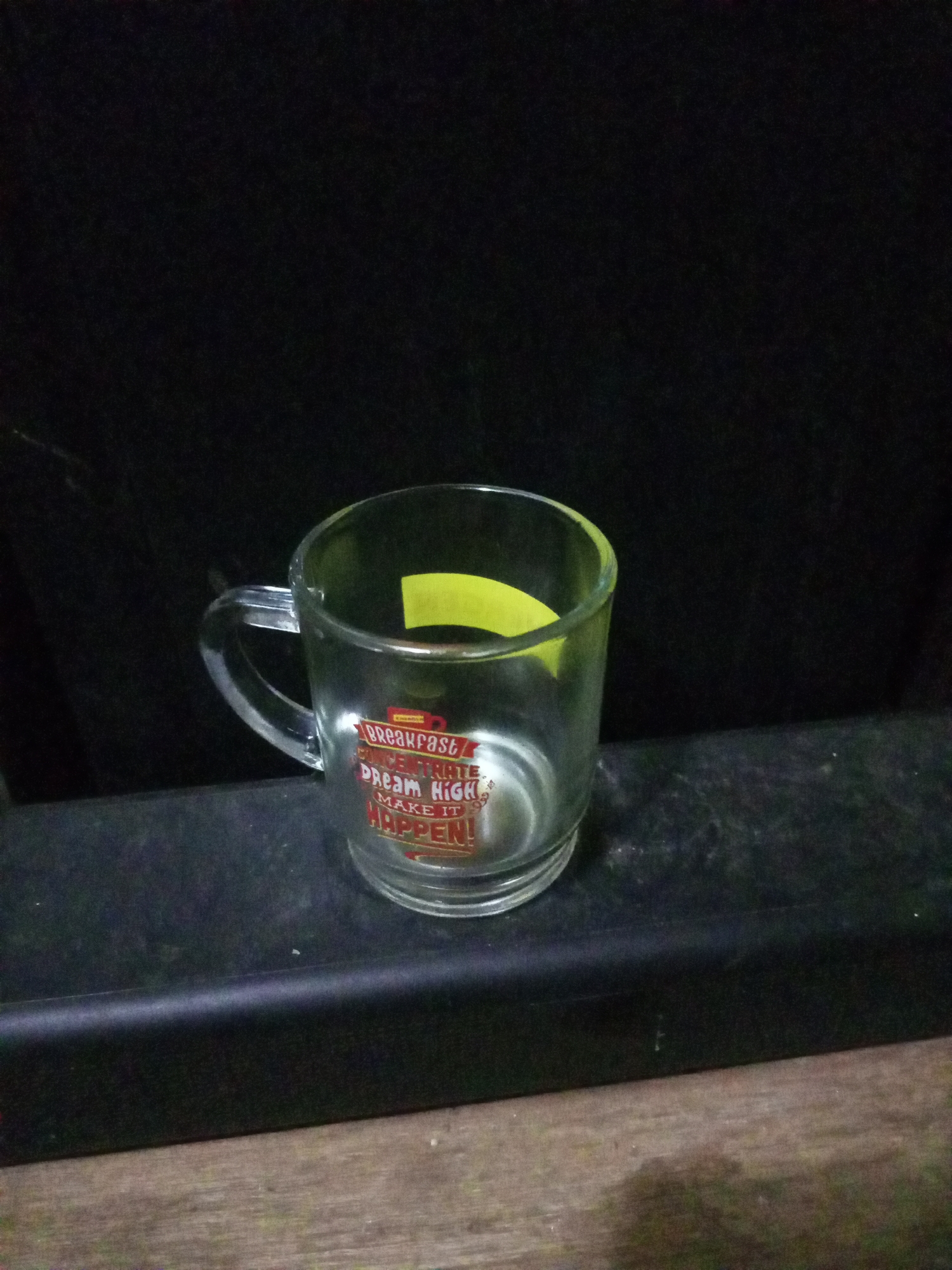 A clear glass mug with a handle rests on a dark, textured surface. The mug has red and white text printed on it that reads "BREAKFAST" and "DREAM HIGH MAKE IT HAPPEN!". A small yellow rectangular sticker is visible on the inside of the mug. The background is dark and out of focus. The surface the mug is on appears to be outdoors on what looks like a wooden table or bench, possibly in a rural setting like Cidahu, Indonesia, given the environment's naturalistic and slightly worn appearance. The lighting is dim, suggesting it might be evening or a shaded area. No people are present in the image.