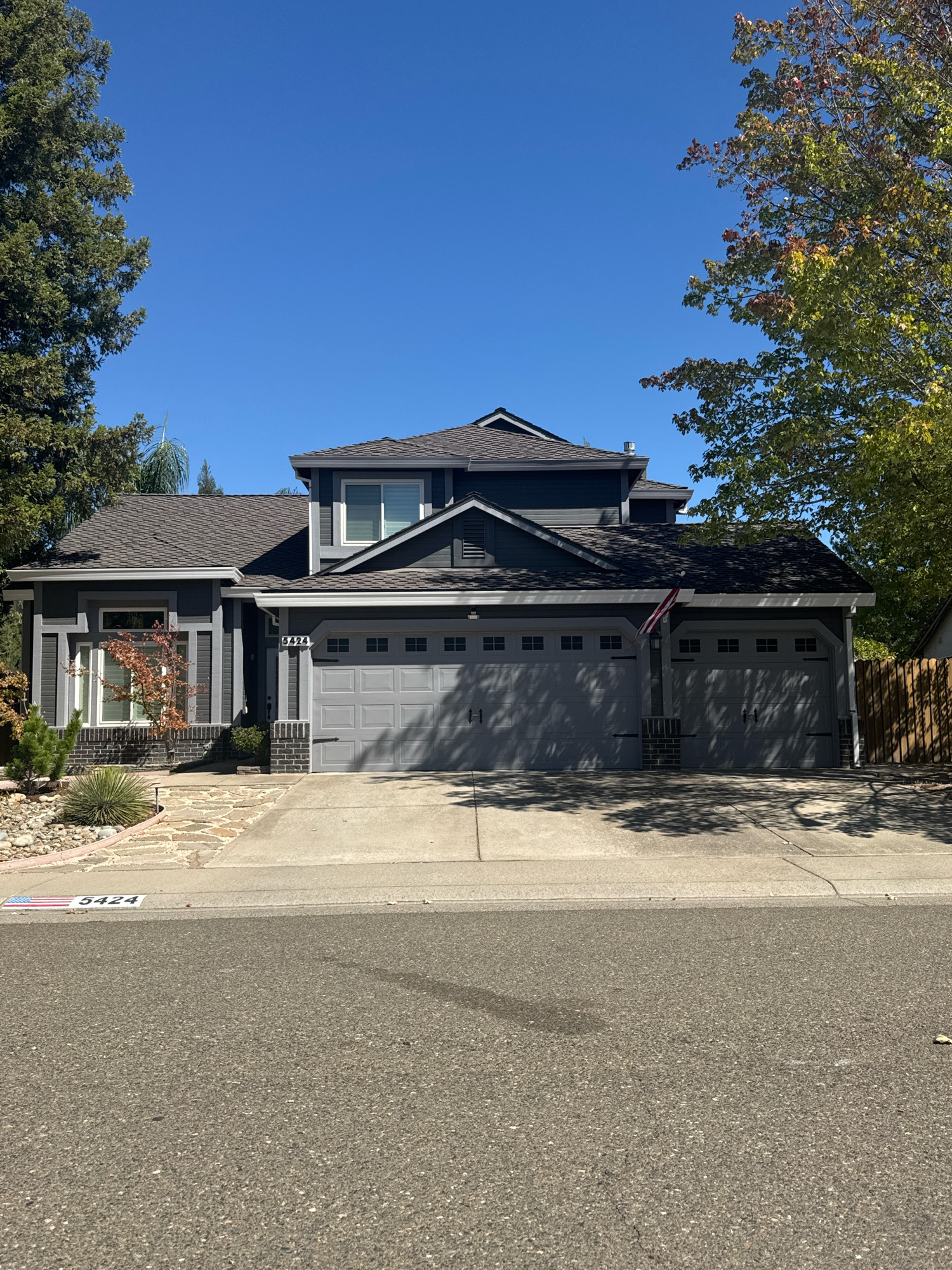 A two-story, dark gray house with a dark gray roof is pictured in Antelope, California, USA. The house features a three-car garage with gray doors and a small American flag attached to a pole in front of the garage. The house number "5424" is visible above the garage. The front yard has a paved driveway, stone pathway, and landscaping with rocks and a small tree. Trees with green and red leaves frame the house, suggesting it is autumn. The sky is clear and blue.