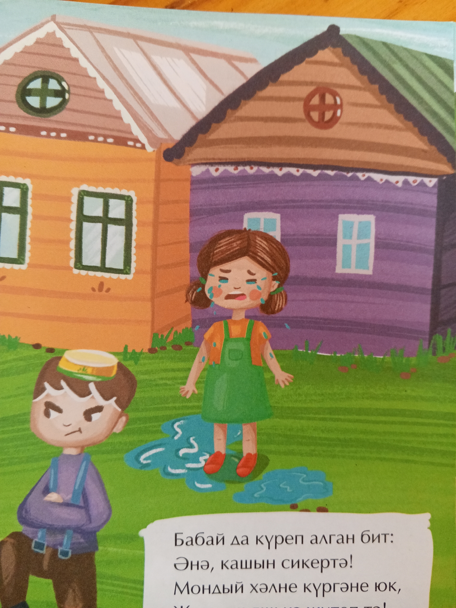 The image depicts a colorful illustration of two cartoon children and two houses. On the left, a boy with brown hair, wearing a purple shirt and brown pants with blue suspenders, stands with his arms crossed and a grumpy expression. He has a yellow and green hat on his head. To his right, a crying girl with brown hair tied in pigtails, wearing an orange shirt and a green overall dress, stands in a puddle of water. She has blue tears streaming down her face and arms. Behind the children are two houses: a yellow house with a light brown roof and a purple house with a green roof. Both houses have windows and decorative trim. The scene is set on a green grassy area. Text is visible at the bottom of the image, but its content is not discernible. There is no indication or visual information within the image that suggests a specific location in Chistopol, Russia, or any other city.