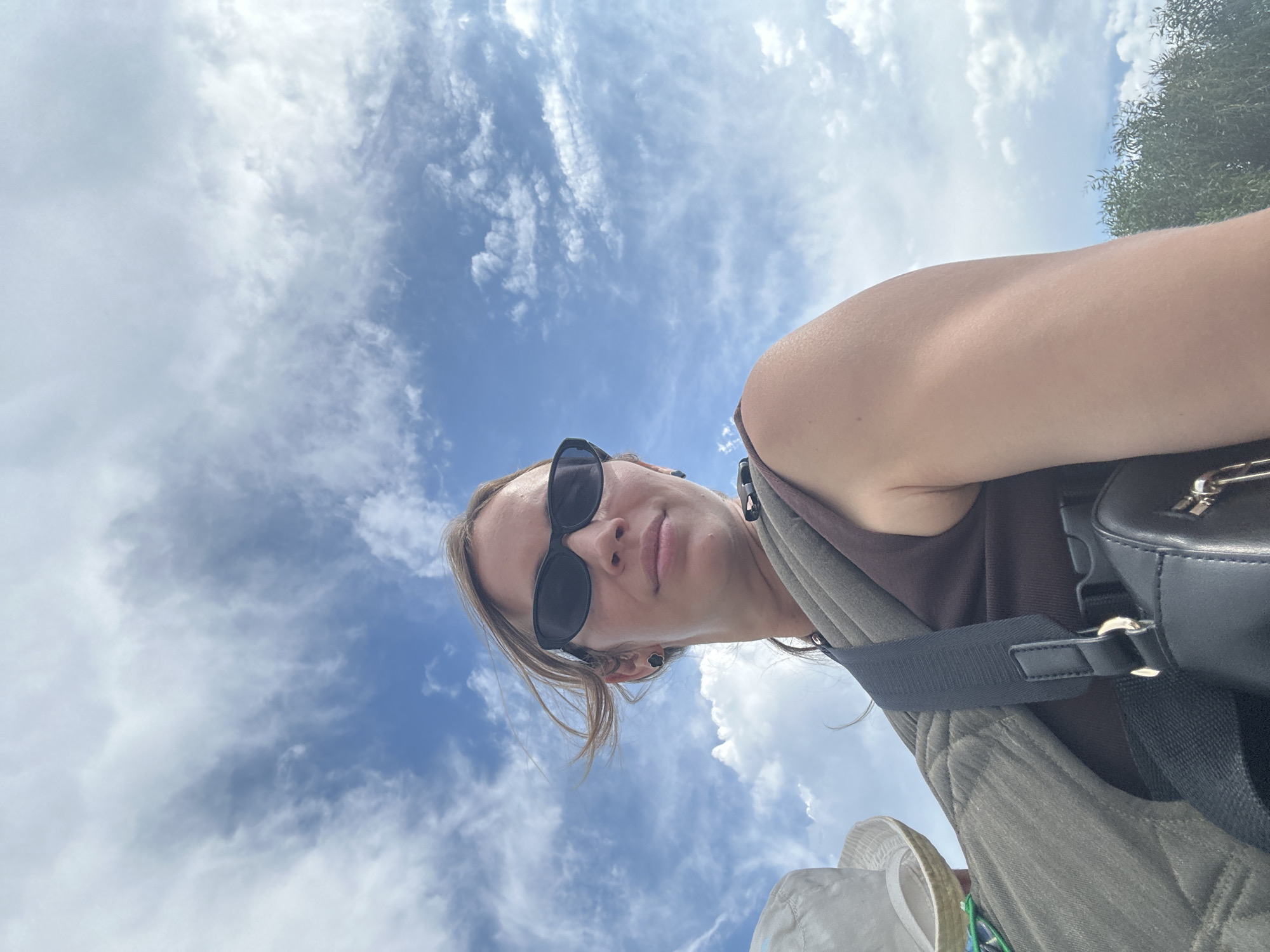 A low-angle, upward-facing shot captures a person wearing sunglasses, a brown tank top, and an olive-green vest. A black bag with a shoulder strap is visible on their right side. The person's blonde hair is pulled back. Behind the person is a blue sky filled with fluffy white clouds. At the top right edge of the frame, the green foliage of a tree is seen. The location appears to be outdoors during daytime, possibly in the city of Fethiye, Turkey.